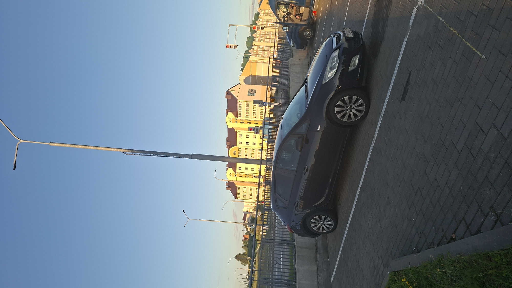 A black Opel Astra is parked in a paved parking lot. The car is facing right and is the main subject of the image. The parking lot is made of dark grey interlocking bricks. A white line marks the parking space. Beyond the parking lot, there is a metal fence, and behind that, a road with traffic lights and a row of yellow and orange apartment buildings. The sky is clear and blue. The lighting suggests it is either early morning or late afternoon. The location is identified as Smaliavichy, Belarus.