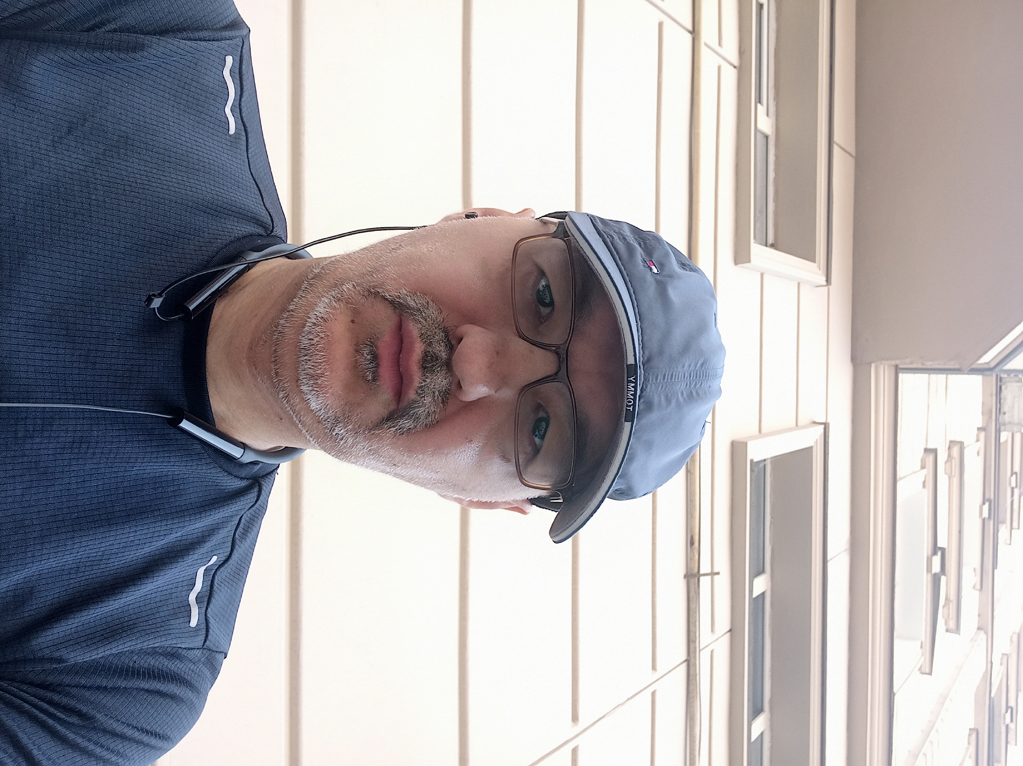 A middle-aged man with light skin, a mustache, and a goatee is captured in a selfie. He is wearing a dark blue baseball cap with a small logo on the front, dark-rimmed glasses, and a dark blue long-sleeved athletic shirt. A pair of black earphones with a connecting band around his neck are visible. The man is looking directly at the camera.
The background consists of the facade of a building with horizontal beige siding and recessed windows. The perspective suggests the photo was taken from a low angle, looking upwards.
This image does not provide any specific visual cues to determine its location within the city of Dushanbe, Tajikistan.