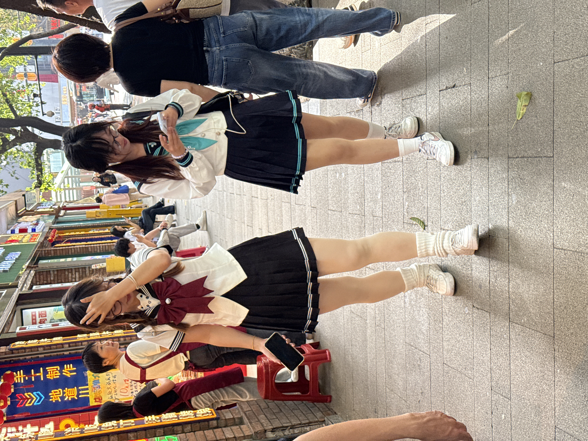 This image captures a vibrant street scene, likely in a commercial area, during bright daylight. The ground is paved with light grey square tiles, and strong sunlight casts distinct shadows.
The primary subjects are two young women in "JK uniform" style outfits, positioned in the foreground and mid-ground. The woman in the mid-ground wears a white long-sleeved top with a teal sailor collar and matching ribbon, a dark pleated mini-skirt with teal stripes, white ankle socks, and white sneakers. She is looking down at a white smartphone she holds in her hands. Her long dark hair flows over her shoulders, and she wears glasses.
Further back and to the right, another woman, partially visible, is wearing jeans and a black short-sleeved top, carrying a woven bag. Her back is to the viewer, and she appears to be walking.
In the foreground, another young woman in a similar school-girl style outfit is seated on a red plastic stool. She wears a white top featuring a large maroon bow at the neckline, and a dark pleated mini-skirt with subtle stripes of white, maroon, and orange. She holds a black smartphone in one hand and has her other hand to her forehead, possibly shielding her eyes from the sun or adjusting her hair. She also wears white ankle socks and white sneakers.
The background reveals a bustling street with several people, some walking, others sitting. Shopfronts line the left side of the street, adorned with various Chinese signs, many of which are neon. A prominent blue neon sign displays large Chinese characters, including "重庆" (Chóngqìng) and "洪崖洞" (Hóngyádòng), indicating a specific location in China, consistent with the provided context of Yuzhong District. Trees are visible on the left, contributing to the outdoor setting. A single withered leaf lies on the pavement in the mid-ground. The overall impression is a casual, lively day in a commercial district.