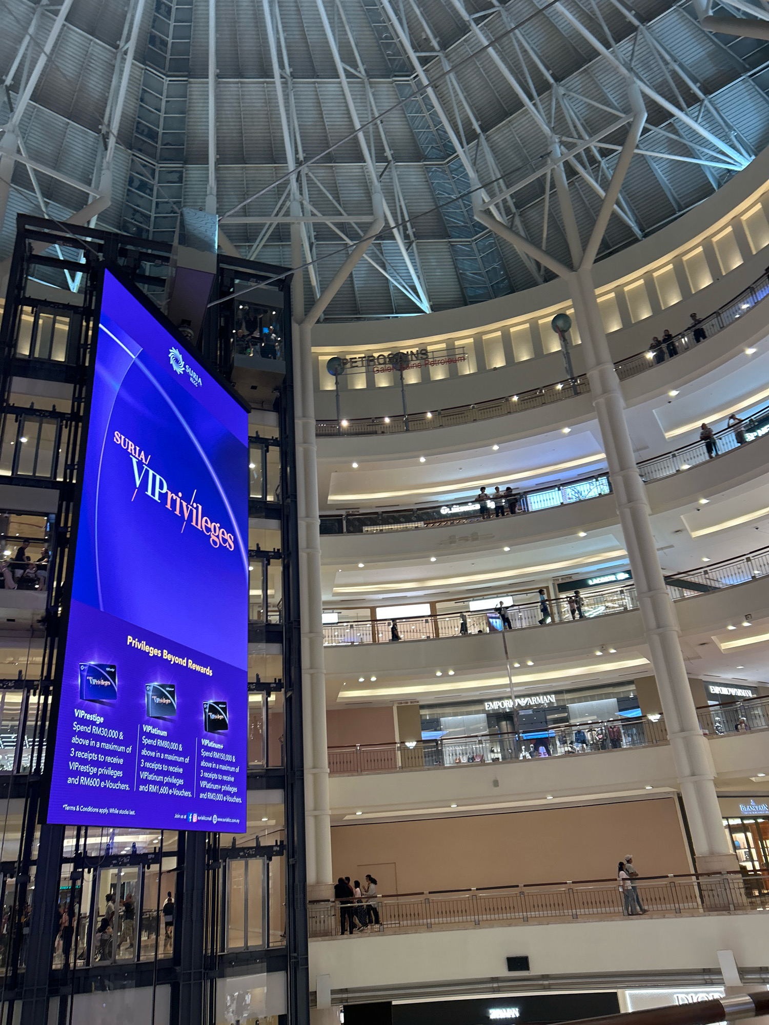 The image depicts the interior of Suria KLCC, a prominent shopping mall located at the base of the Petronas Twin Towers in Kuala Lumpur, Malaysia. A large, illuminated blue digital screen displays information about "SURIA VIPrivileges," outlining different tiers like VIPrestige, VIPatinum, and VIPatinum+ with associated spending requirements and rewards in e-vouchers. The screen hangs from a complex, multi-level atrium structure with a domed, translucent ceiling supported by white steel beams. Several floors of the mall are visible, featuring escalators, railings, and storefronts. People are seen on various levels, some walking along the balconies, others inside shops, and a few gathered near the digital display. The environment is bright and modern, characteristic of a large, upscale shopping complex. The iconic architecture of the mall's interior, with its expansive scale and layered levels, is a key feature of the scene.