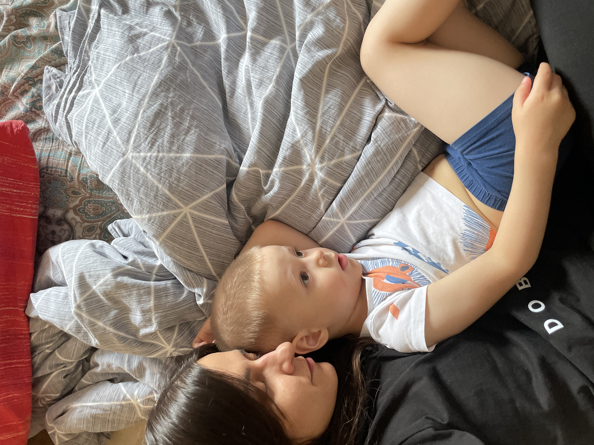 An overhead shot captures a woman and a young child lying side-by-side on a bed, sharing a quiet moment. The woman, with dark hair, is positioned on the right side of the frame, wearing a black top with partially visible white letters that read "DOB". She is looking gently towards the child. The child, with light blonde hair, lies next to her, wearing a white t-shirt with orange and blue graphic designs and blue shorts. The child's gaze is directed upwards and slightly to the side, with an alert and curious expression.
The bed is covered with varied bedding: a light gray duvet with a prominent white geometric pattern of intersecting lines, and a section of sheets featuring a blue and brown paisley-like design. A red pillow or cushion is partially visible along the left edge of the frame. The lighting suggests daytime, with soft, diffused light illuminating the scene, creating a sense of calm and intimacy. The activity appears to be resting or relaxed interaction between the two individuals in what is likely a bedroom setting.