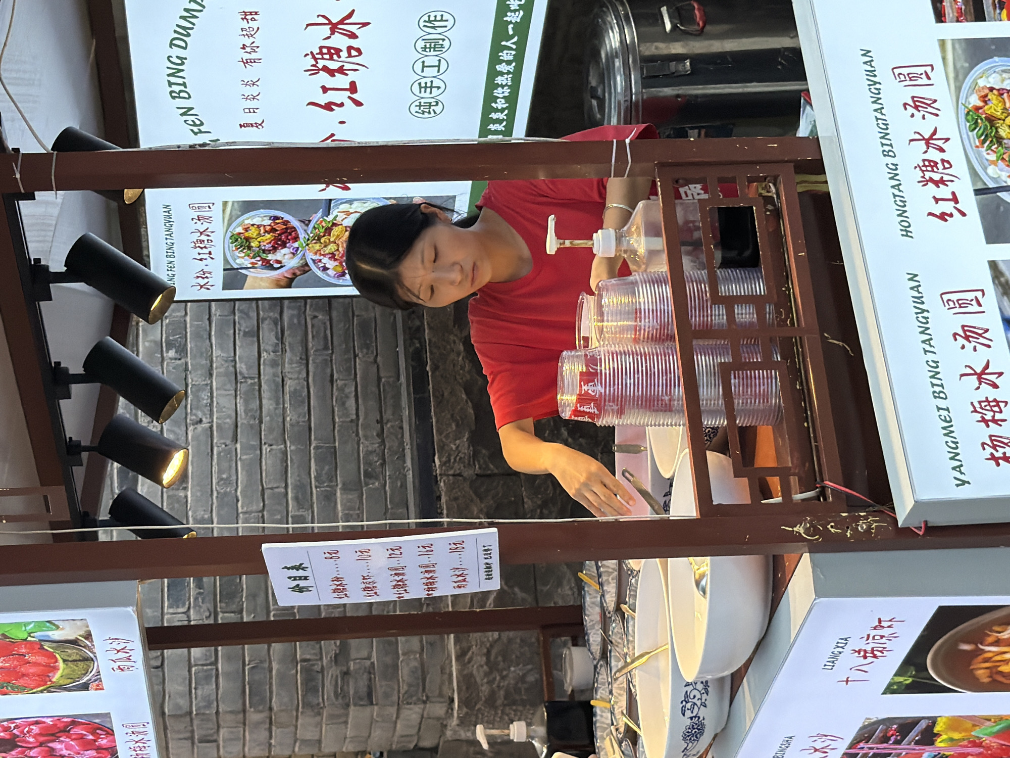In Yuzhong District, China, a young woman in a red t-shirt operates a food stall. The stall, constructed of dark wood, displays various food items and prices on white signs. In the foreground, several large white bowls filled with what appears to be a creamy liquid and spoons are arranged, suggesting prepared desserts or drinks. To the right of the bowls, stacks of clear plastic cups are visible, along with a pump dispenser. The woman is actively engaged in her work, her hands near the cups and a ladle. In the background, a brick wall forms the backdrop, and several modern black track lights illuminate the stall. Signs above the stall display Chinese characters and images of food, with one sign featuring photos of a fruit dessert. Another sign on the left side of the stall includes a list of prices for various items like "Red Bean Soup," "Red Bean Paste," and "Tangyuan." The overall scene depicts a bustling street food vendor in a commercial area.