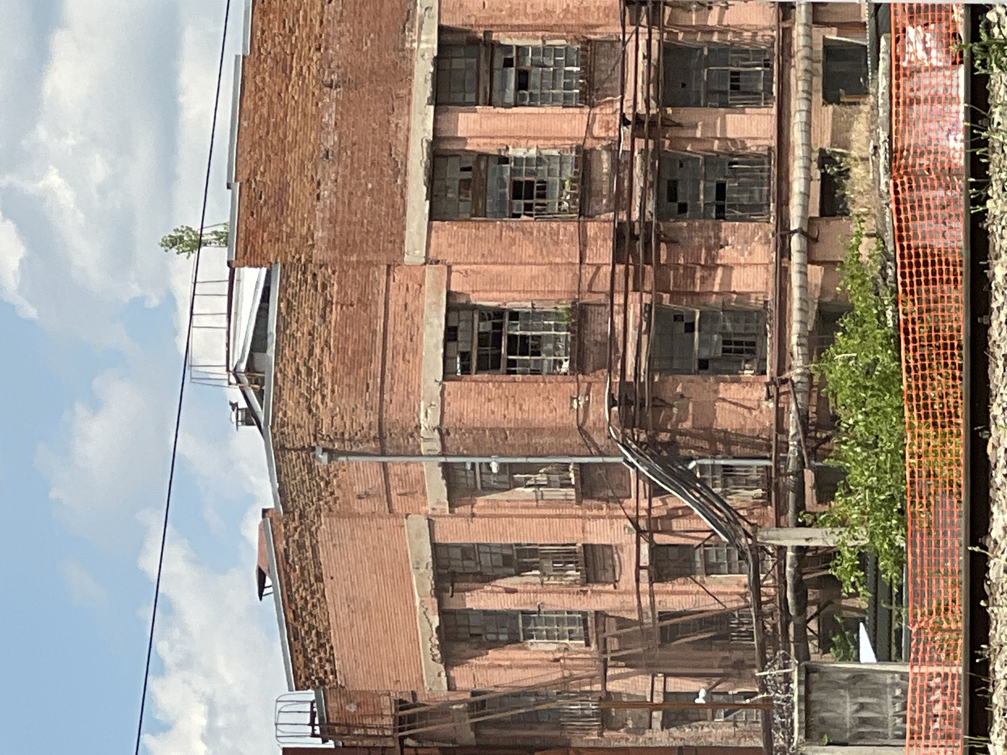 An exterior, medium-range, eye-level shot captures a weathered, pink brick industrial building under a partly cloudy blue sky. The building exhibits signs of decay; many windowpanes are broken or missing, and sections of the brickwork appear eroded. Various metal structures, including pipes, scaffolding, and what appears to be a fire escape, are attached to the facade. Dense green foliage sprouts near the base of the building, partially obscured by an orange mesh fence. Railway tracks and ballast are visible in the foreground. No people are present in the image. The building is located in the city of Krasnodar, Russia, specifically at the site of the former Krasnodar Brick Factory No. 2, now part of the Krasnodar Construction Materials Plant.