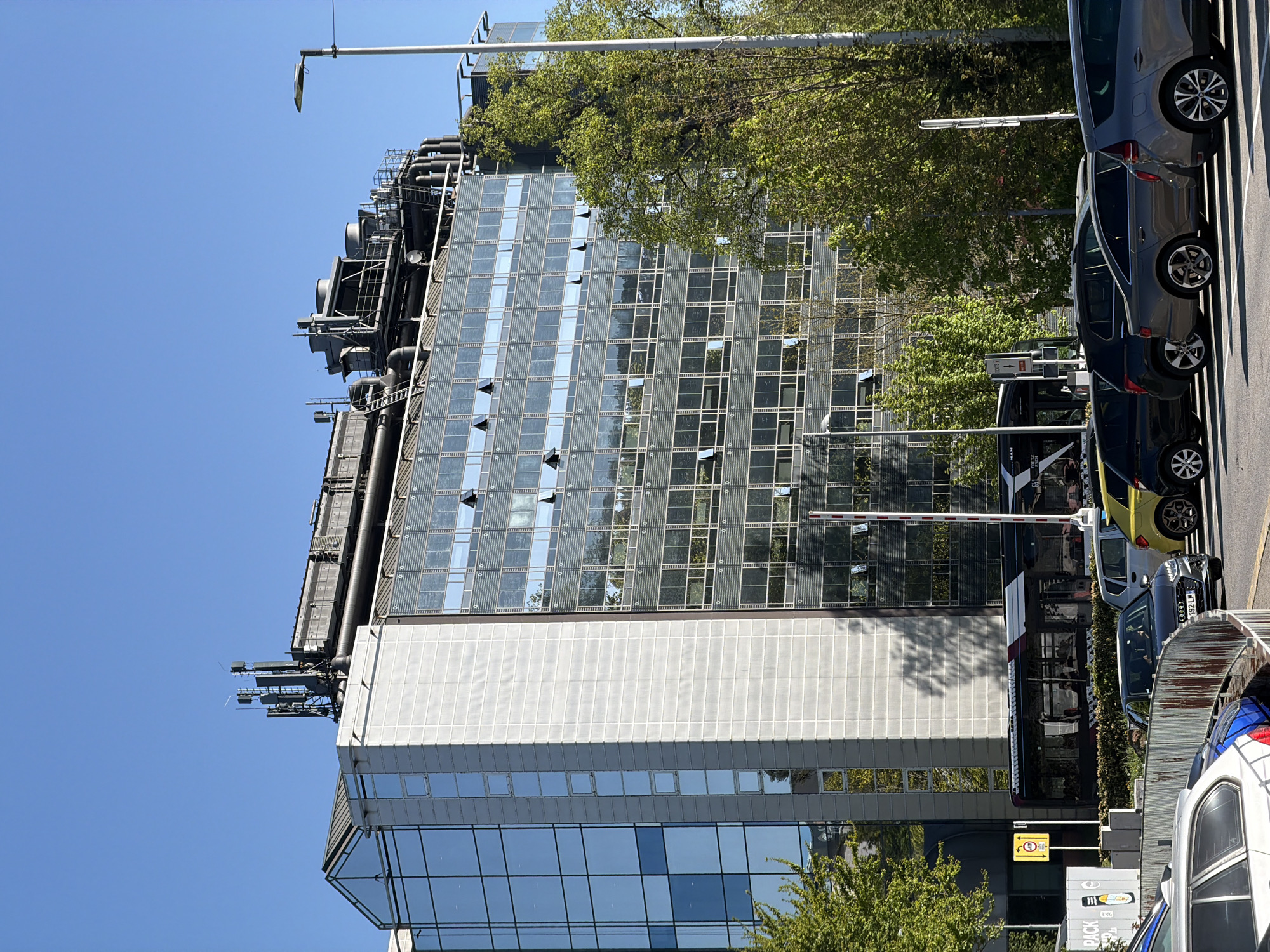 The image captures a modern building in Esch-sur-Alzette, Luxembourg, under a clear blue sky. The building features a multi-level design with a combination of reflective glass panels, white concrete sections, and rooftop mechanical systems. Several windows are partially open. The surrounding environment includes trees with green foliage, a light pole, and parked cars along the adjacent street. A bus is seen at a bus stop. The photograph is taken from a ground-level perspective.