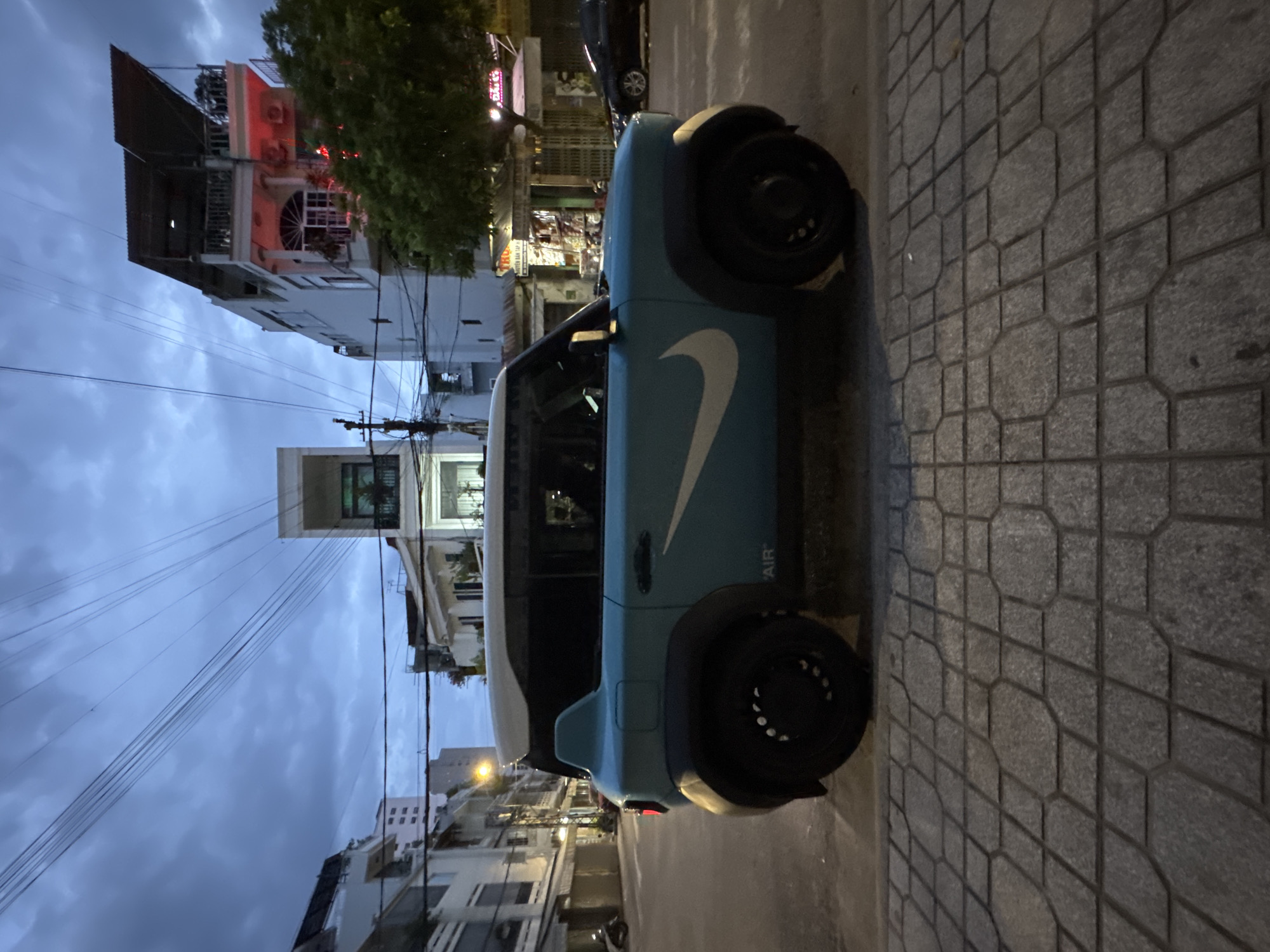 A turquoise and white compact car, customized with a large white Nike swoosh logo and the word "AIR" in small black lettering on the side, is parked on a sidewalk. The car is positioned on a street in what appears to be a bustling urban environment, likely in Nha Trang, Vietnam, given the architectural style of the surrounding buildings, which include multi-story structures with balconies and outdoor living spaces, as well as storefronts. The sky is overcast and twilight is setting in, casting a dim, cool light on the scene. The sidewalk is paved with hexagonal and rectangular stone tiles. In the background, other vehicles, including a black car and a motorcycle, are visible on the street, and overhead power lines crisscross the sky.