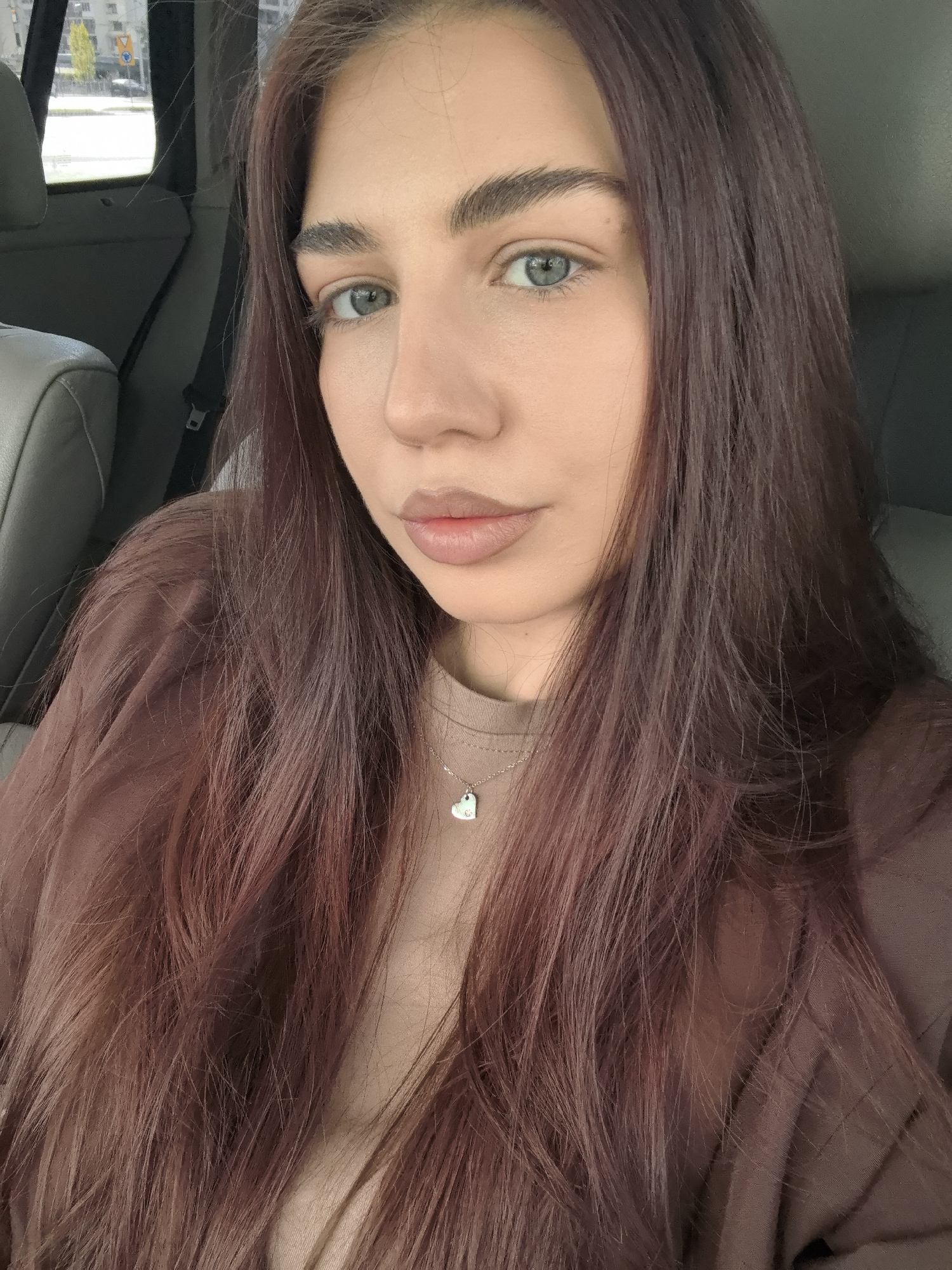 A close-up selfie shows a young woman with long, reddish-brown hair sitting in a car. She has light skin, green eyes, and full lips. She is wearing a tan t-shirt and a brown overshirt. A silver necklace with a heart pendant is visible. The car's gray interior is visible in the background. Through the car window, an urban environment is suggested, potentially depicting a section of Stara Zagora, Bulgaria, based on the request. The scene appears to be taken during daylight hours. The woman is looking directly at the camera.