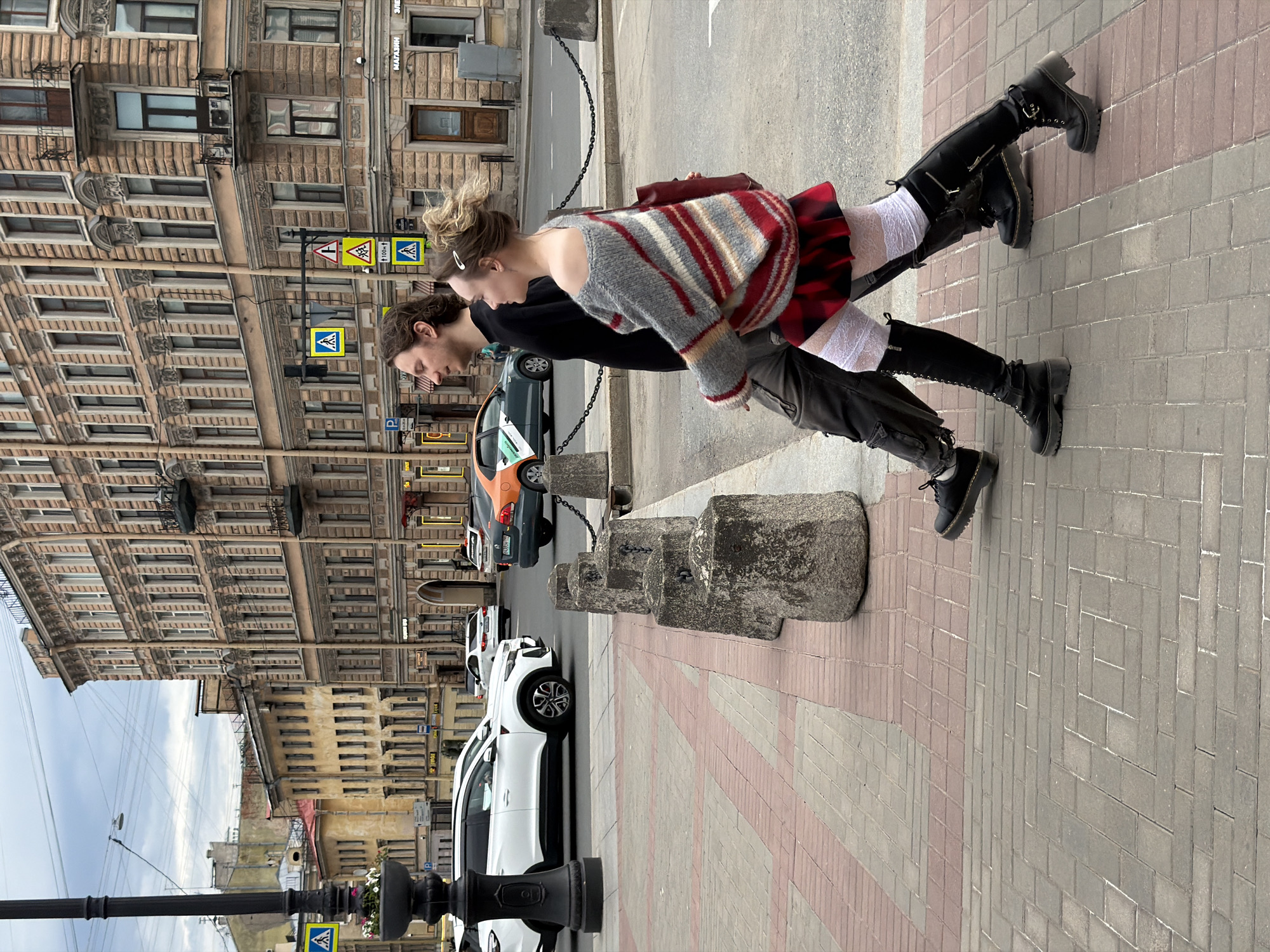 This image captures an urban street scene in Saint Petersburg, Russia, characterized by its distinctive architecture and pedestrian activity.
In the foreground, two individuals are walking from right to left on a paved sidewalk. The person on the left is a male with medium-length, light brown wavy hair, a light beard, wearing a black long-sleeved shirt, dark cargo pants, and black shoes. He is looking forward with a slight smile. The person on the right is a female with blonde hair styled in an updo, secured by a white clip. She is wearing an off-the-shoulder grey, red, and white striped sweater, a red and black plaid pleated mini-skirt, white lace-patterned over-the-knee stockings, and black knee-high lace-up boots with thick soles and heels. She carries a dark red shoulder bag on her right side and is looking slightly forward and to her left. Both appear to be engaged in a casual walk.
The sidewalk is paved with rectangular bricks arranged in alternating patterns of grey/beige and reddish-brown sections. A line of dark grey, octagonal stone bollards linked by a metal chain separates the sidewalk from the asphalt street. On the far left, a black lamppost with a decorative base features a planter containing white flowers.
The street accommodates vehicular traffic. A white SUV is partially visible on the left, and a dark green car with an orange decal is positioned behind the bollards. Further down the street, a white sedan and an orange and dark grey taxi are visible. Overhead, an overcast sky is present.
The background is dominated by multi-story buildings. On the right, a large, ornate building with a light reddish-brown facade features neoclassical architectural elements, including decorative window frames, cornices, pilasters, and multiple balconies. The ground floor displays shopfronts, one with "МАГАЗИН" (Shop) and "ЭЛЕК..." (Elec...) visible in Cyrillic lettering. Further down the street on the left, an older, yellowish multi-story building with simpler architectural details is visible. Several blue square pedestrian crossing signs are present, one on the lamppost and another above the walking individuals, along with a triangular warning sign indicating a pedestrian crossing and a rectangular sign marking "100m." A blue "P" parking sign is also visible on the left side of the street.