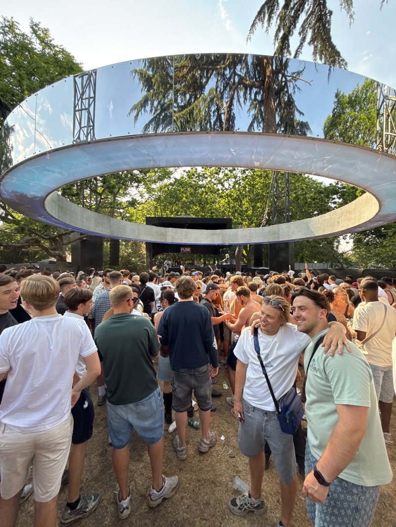 A large crowd of predominantly young people, likely festival-goers, are gathered in an outdoor park setting. Above them is a large circular structure, potentially part of a stage design or art installation, with a mirrored or reflective exterior. The crowd is dense, with people standing, talking, and interacting. The ground appears to be grass or dirt, possibly compacted from foot traffic. The backdrop includes trees and possibly part of a stage setup. The image does not provide explicit evidence to confirm the location is in Barking, United Kingdom. Further context would be needed to definitively establish the city of Barking as the location.