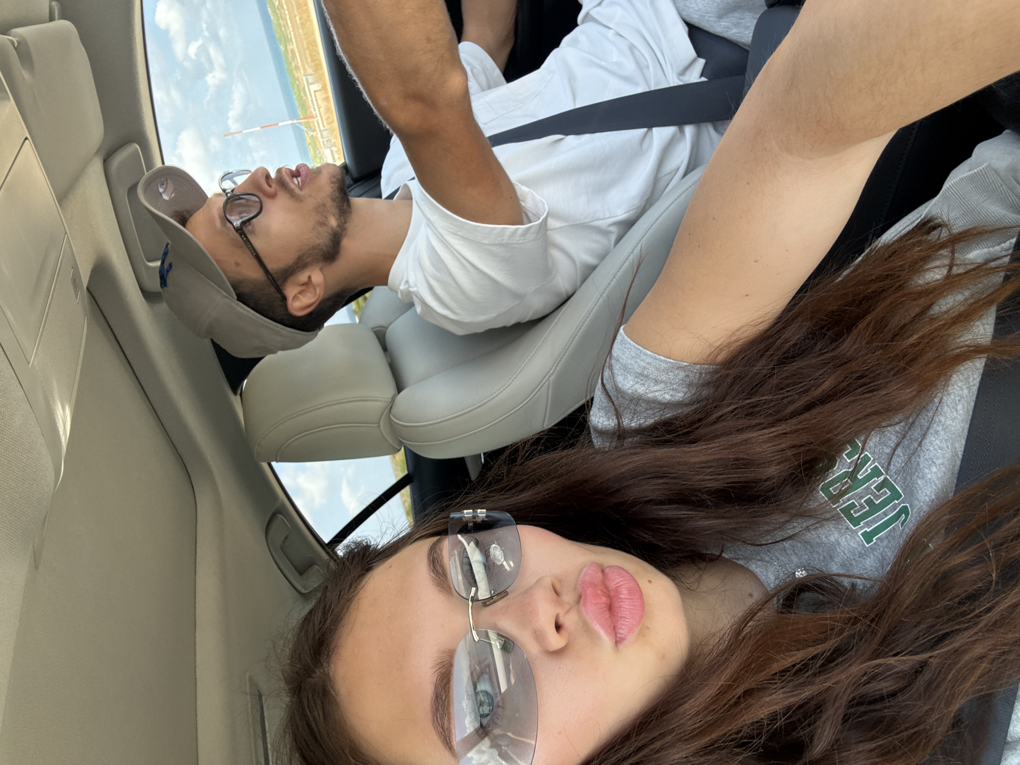 Inside a vehicle, a woman with long brown hair, sunglasses, and a grey t-shirt occupies the foreground, while a man wearing glasses, a cap, and a white shirt is in the background. Both appear to be wearing seatbelts. Outside the vehicle, a partial view of the landscape including a pale sky and a structure resembling a tower or mast can be seen. The picture was taken within a vehicle in Hof HaCarmel Regional Council, Israel.