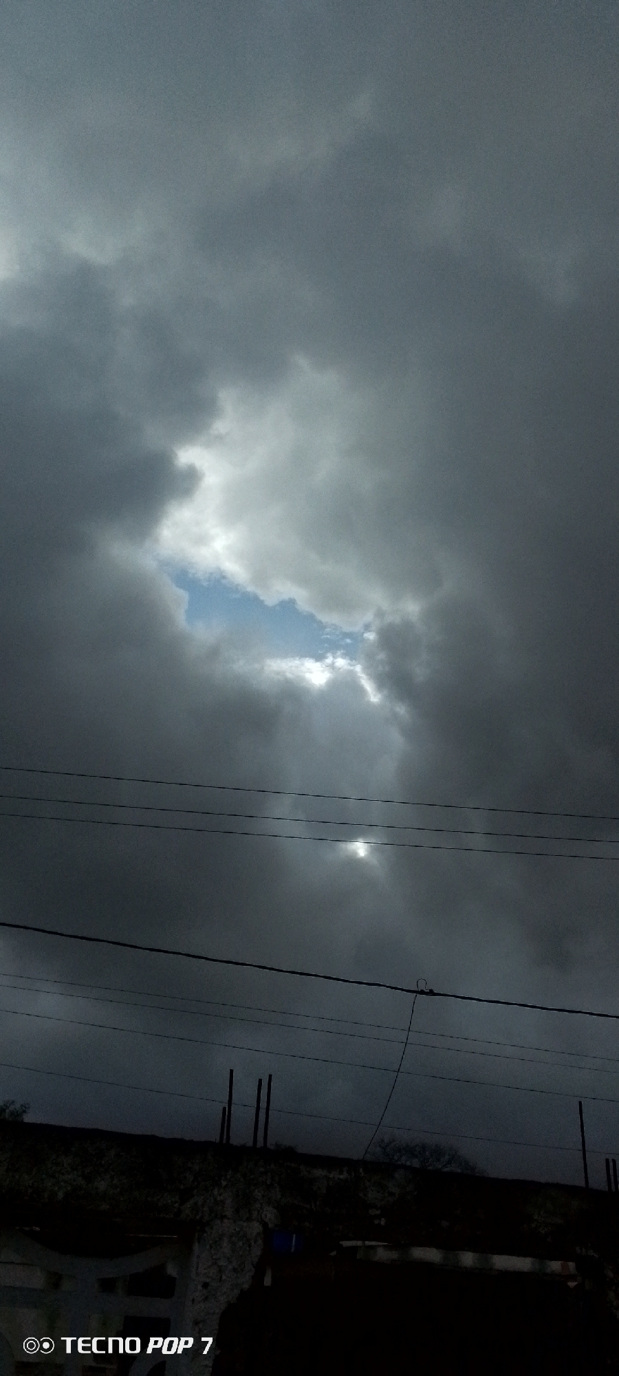 The image depicts a dramatic sky over Altagracia, Venezuela. Heavy, dark storm clouds dominate the upper portion of the frame, with a break in the clouds revealing a patch of bright blue sky and the sun peeking through, casting visible rays. Several horizontal power lines stretch across the middle ground. The lower part of the image shows a dark, silhouetted landscape that appears to be rooftops or elevated structures with some vertical elements suggesting utility poles or unfinished construction. The bottom left corner includes a watermark that reads "TECNO POP 7". There are no people or animals visible in the scene.