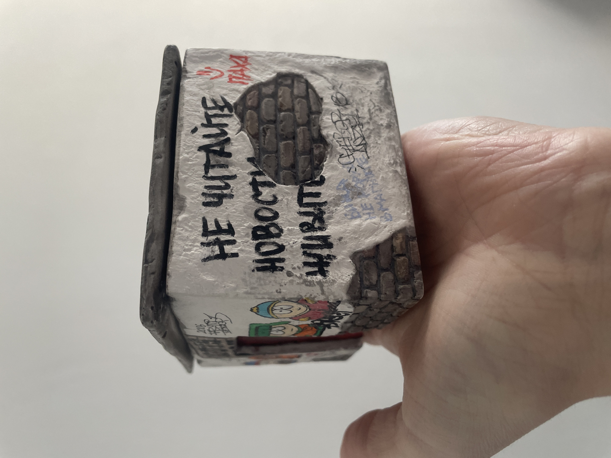 A hand holds a small, rectangular object resembling a building or structure. The object is designed to look weathered, with faux brickwork visible in some areas and a painted white surface that appears to be aged or decayed. Graffiti-like markings and drawings cover parts of the object, including the inscription "НЕ ЧИТАЙТЕ НОВОСТИ ЖИВИТЕ" (Do not read news, live). The object also features cartoonish drawings of characters that resemble South Park figures. The scene is against a plain, white background.