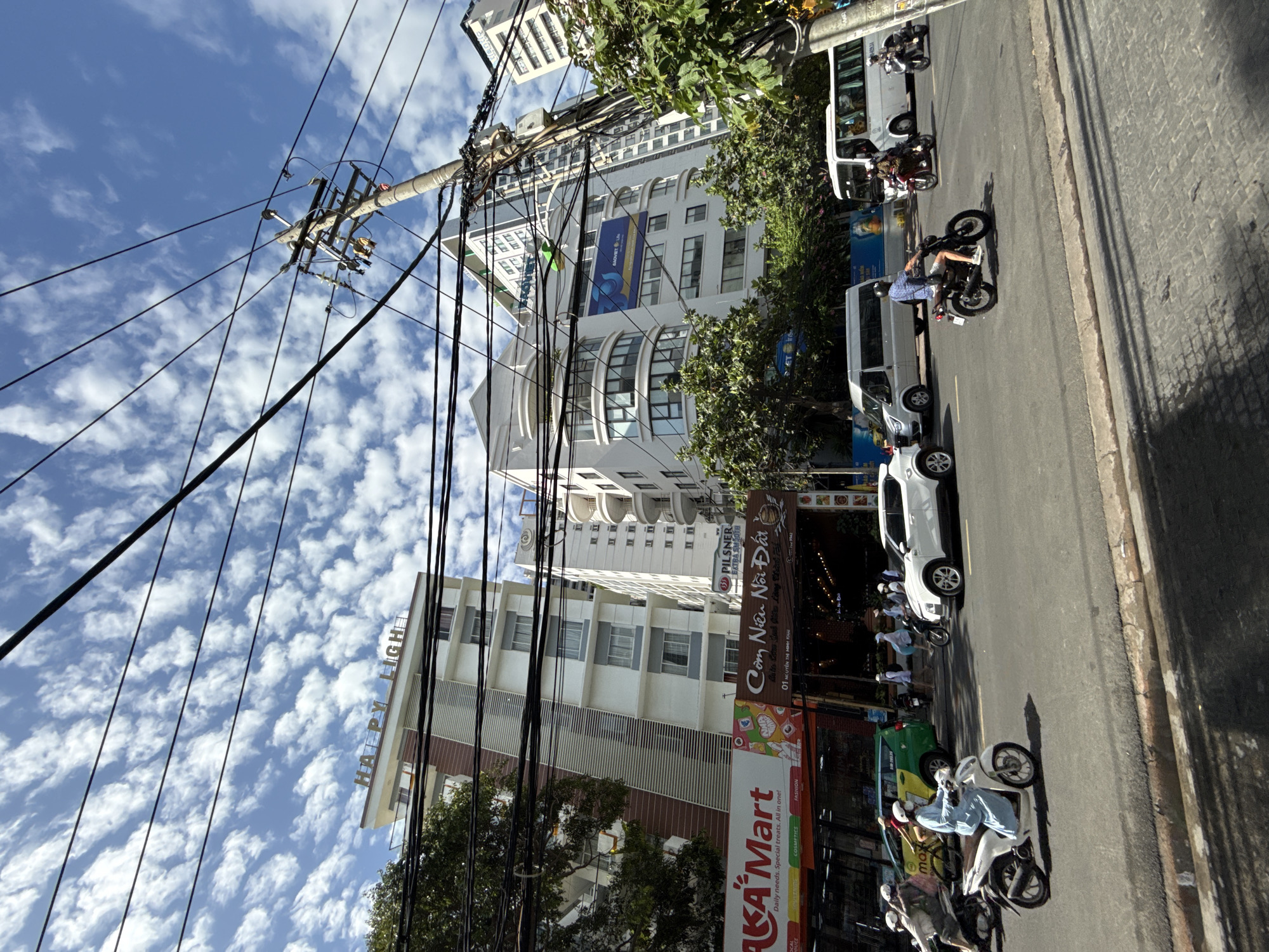 The image depicts a street scene in Nha Trang, Vietnam, under a bright, cloudy sky. Several buildings line the street, with one prominently featuring the text "HAPPY LIGHT" on its facade. Various types of vehicles, including motorcycles, cars, and vans, are present on the asphalt road. People are seen riding motorcycles, walking, and some are wearing face masks. Overhead, a complex network of electrical wires crisscrosses the sky, attached to a utility pole. Businesses such as "AKA Mart" and "Pilsner" are visible on the lower floors of some buildings. Trees and foliage add to the urban landscape. The overall impression is one of a bustling city street during daylight hours.