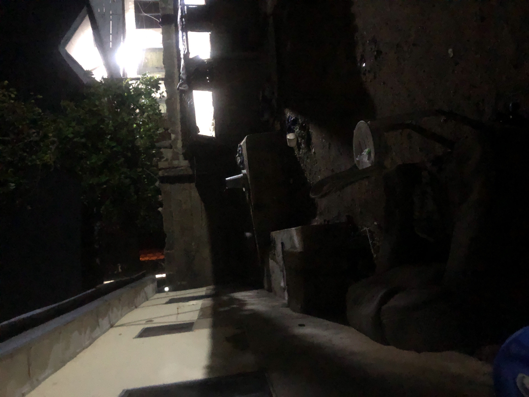 The image depicts an outdoor scene at night, illuminated by artificial light sources, suggesting an urban environment in Kabong, Nigeria. A building with a light-colored exterior and dark windows is visible on the left side of the frame, extending into the background. Adjacent to it, a brick wall separates the property from an area with rubble and debris. In the mid-ground, there are concrete structures, possibly remnants of old construction, and a plastic chair with a white plate on it. Further back, a house with a brightly lit interior and a visible roofline stands out. A large tree with dark green foliage is situated between the building on the left and the house in the background. The ground is uneven and appears to be covered with dirt, scattered leaves, and possibly trash. The overall impression is one of a residential area, potentially with ongoing construction or in a state of disrepair, as indicated by the debris and unfinished structures. The lighting suggests it is nighttime, and the shadows cast by the artificial lights create a somber atmosphere.