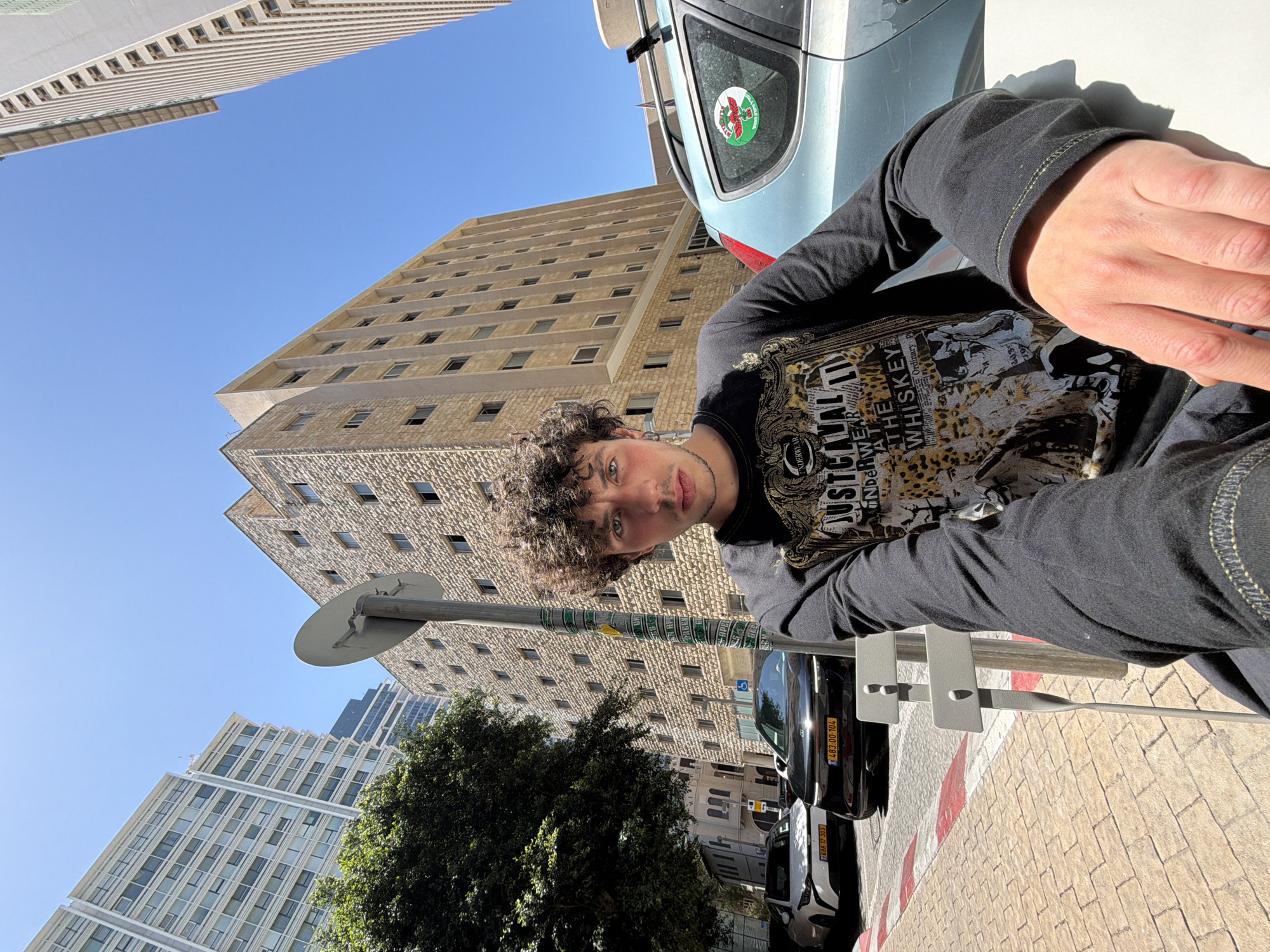 A tilted perspective of a young man stands in Tel Aviv, Israel, with curly hair, a black shirt with a graphic design, and fair skin, looking toward the camera. A traffic pole is to his left, and behind him, a beige multi-story building and a blue car are visible. Further back, a tree and another building are present. The sky is clear blue. A white car is parked on the street with two separate license plates. The license plates are marked with "698 17-303" and "483:00-104" respectively.