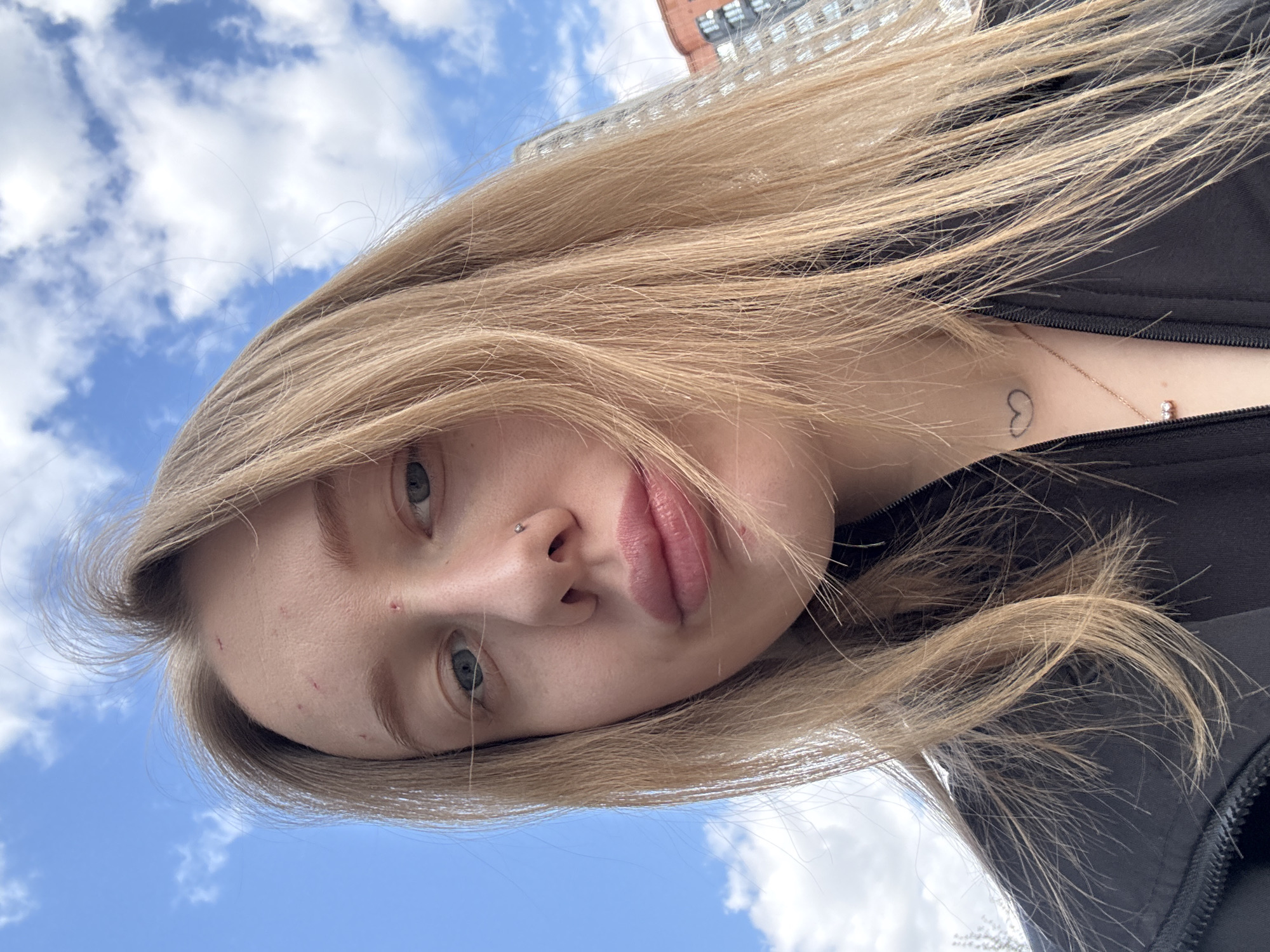 This image is a close-up, tilted portrait of a young woman with long, flowing blonde hair, captured outdoors on a bright day. Her head is angled, showing her face in the lower-left to central part of the frame, looking up and slightly to the right. Her blue eyes are visible, one looking towards the viewer. She has a small silver stud piercing on her left nostril and full, neutral lips.
Her blonde hair, appearing windswept, dominates the upper and right portions of the image, creating a dynamic element. She is wearing a dark-colored garment, possibly a black jacket, visible around her neck and shoulder. A small, outlined heart tattoo is visible on her left collarbone, alongside a delicate gold chain with a small, light-colored pendant. Minor skin imperfections are noticeable on her forehead.
The background features a clear blue sky dotted with scattered white clouds, confirming an outdoor setting during the daytime. In the far upper-right corner, a section of a reddish-brown brick building with windows is visible, suggesting an urban or city environment. The perspective is an upward-looking shot, possibly a selfie. The woman's expression is neutral and calm. There is no visible text in the image.