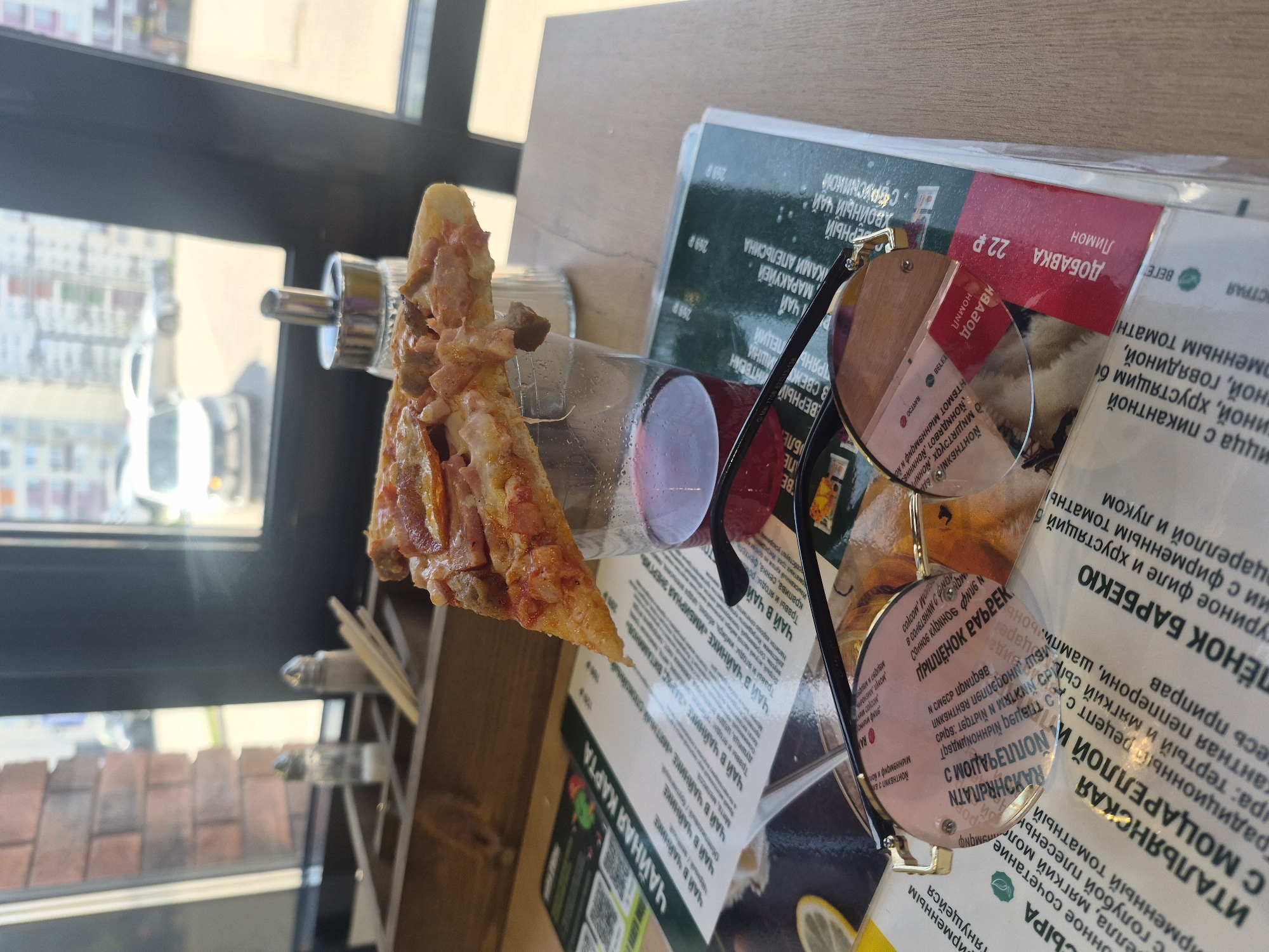 The image depicts a table setting indoors, likely in a restaurant or cafe. A slice of pizza rests on its side, leaning against a clear bottle containing a red liquid. Nearby is a menu with text in Russian, partially obscured by a pair of round eyeglasses with black frames. The location appears to be Izhevsk, Russia, based on the Russian language on the menu. The table is wooden, and outside the window, a cityscape is visible. No people are directly visible. The primary subjects are the pizza, bottle, glasses, and menu. The lighting suggests it is daytime.