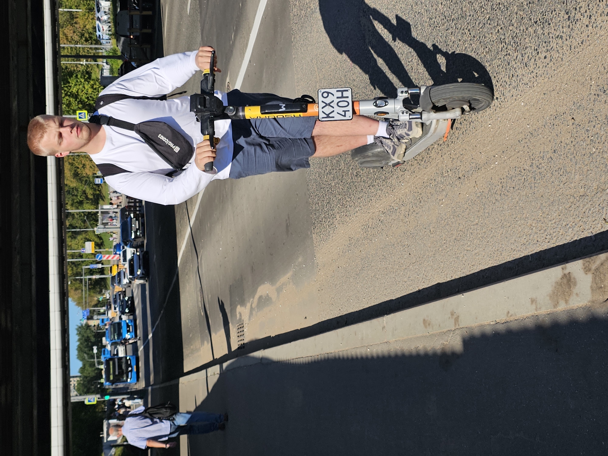 A young blond man is riding an electric scooter down a city street in Moscow, Russia. He is wearing a white long-sleeved shirt, blue shorts, grey sneakers, and white socks. He also has a black fanny pack across his chest. The scooter is gray, silver, and orange, with a license plate that reads "KX9 40H". In the background, there are cars and buses, as well as trees and buildings. A pedestrian wearing a blue plaid shirt and jeans is walking on the sidewalk to the left of the scooter rider. The day is sunny, casting shadows on the road.