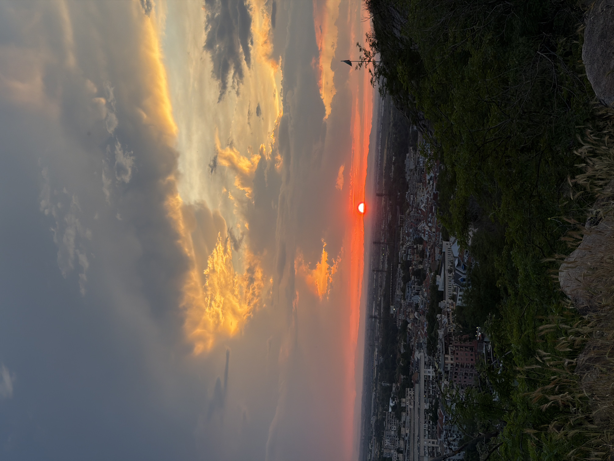 This is a photograph taken from a vantage point on a hill overlooking the city of Plovdiv, Bulgaria, during sunset. The sky is filled with dramatic clouds, illuminated by the setting sun which appears as a bright orange orb near the horizon. The clouds are a mix of dark grays and vibrant oranges and yellows, suggesting a dynamic and colorful sky.
In the middle ground, the city of Plovdiv spreads out, with a dense collection of buildings, some appearing to be residential and others with a more commercial or industrial feel. There are also indications of infrastructure like roads and possibly some towers or masts. The foreground features the natural landscape of the hill, with green foliage, dry grasses, and rocks. A single flag on a pole is visible on the right side of the frame, positioned on the edge of the hill. The overall impression is a scenic view of a city at dusk, with a focus on the natural beauty of the sunset and the surrounding environment.