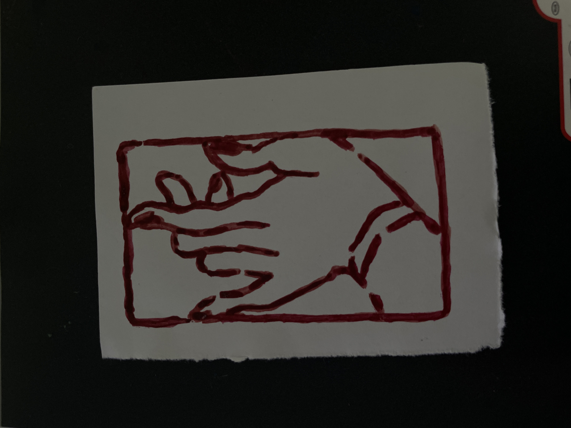 A close-up shot shows a white paper with a red drawing of a hand inside a rectangle. The drawing is stylized and appears to be made with marker or paint. The paper has torn edges on the bottom and left side. The background is dark, suggesting the paper is on a dark surface, possibly a table or floor. The image is taken from a slightly elevated angle. No specific location in Minsk, Belarus can be determined from the visual information provided.