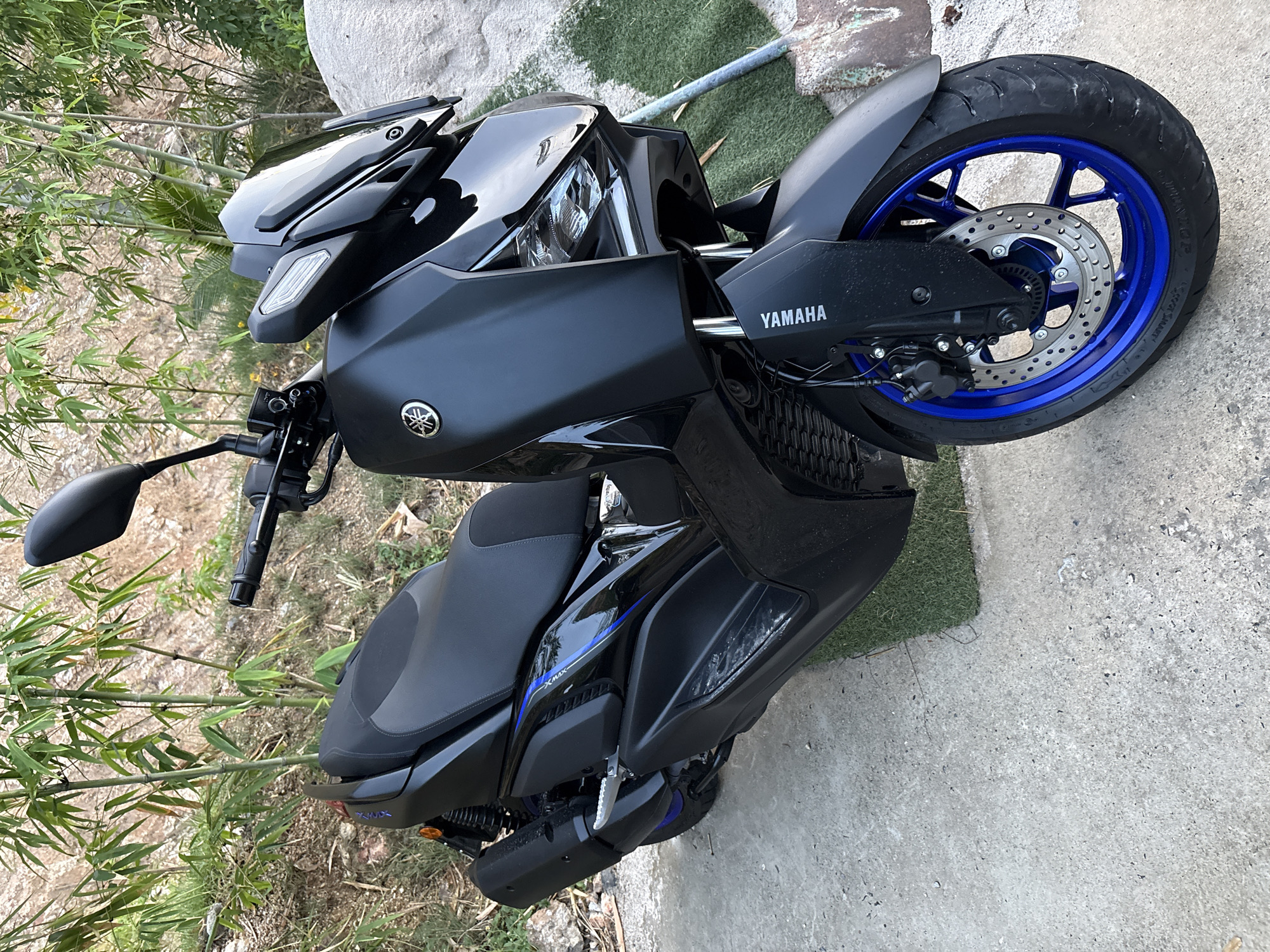 The image shows a black Yamaha motorcycle parked outdoors in Baan Bang Rak, Thailand. The motorcycle is positioned on a concrete surface next to green plants and a large rock. The rear tire features a blue rim. The "YAMAHA" logo is visible on the motorcycle's frame. There are no people visible in the image.
