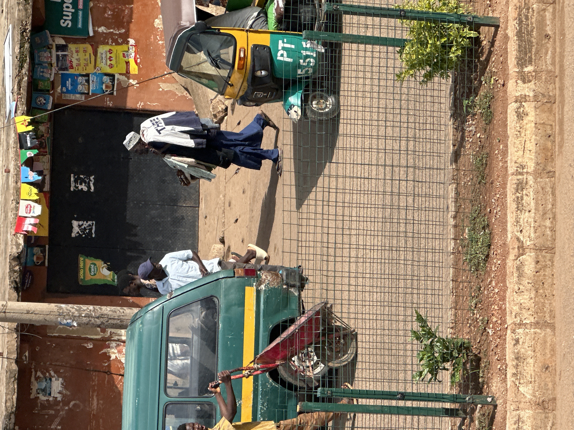 This is a medium shot, eye-level, outdoor photograph taken in Bukuru, Nigeria. The image captures a street scene with several vehicles and people. In the foreground, a green wire fence runs horizontally across the frame, with a small plant growing on the other side. Behind the fence, a green and yellow auto-rickshaw is parked, its license plate reading "PT-5515". To the left of the auto-rickshaw, a green minibus is parked. A man in a yellow t-shirt and brown pants is standing to the left of the minibus, pushing a red wheelbarrow. Another man wearing a blue shirt and a blue cap is standing in front of the minibus. In the background, a building with a brown facade and a black door is visible. There are several posters and signs attached to the building. Two individuals are walking away from the building; one is wearing a blue skirt and a white and dark blue jacket, and the other is wearing a dark blue outfit and a white head covering. The ground is a mixture of dirt and concrete. The scene appears to be a busy urban environment.