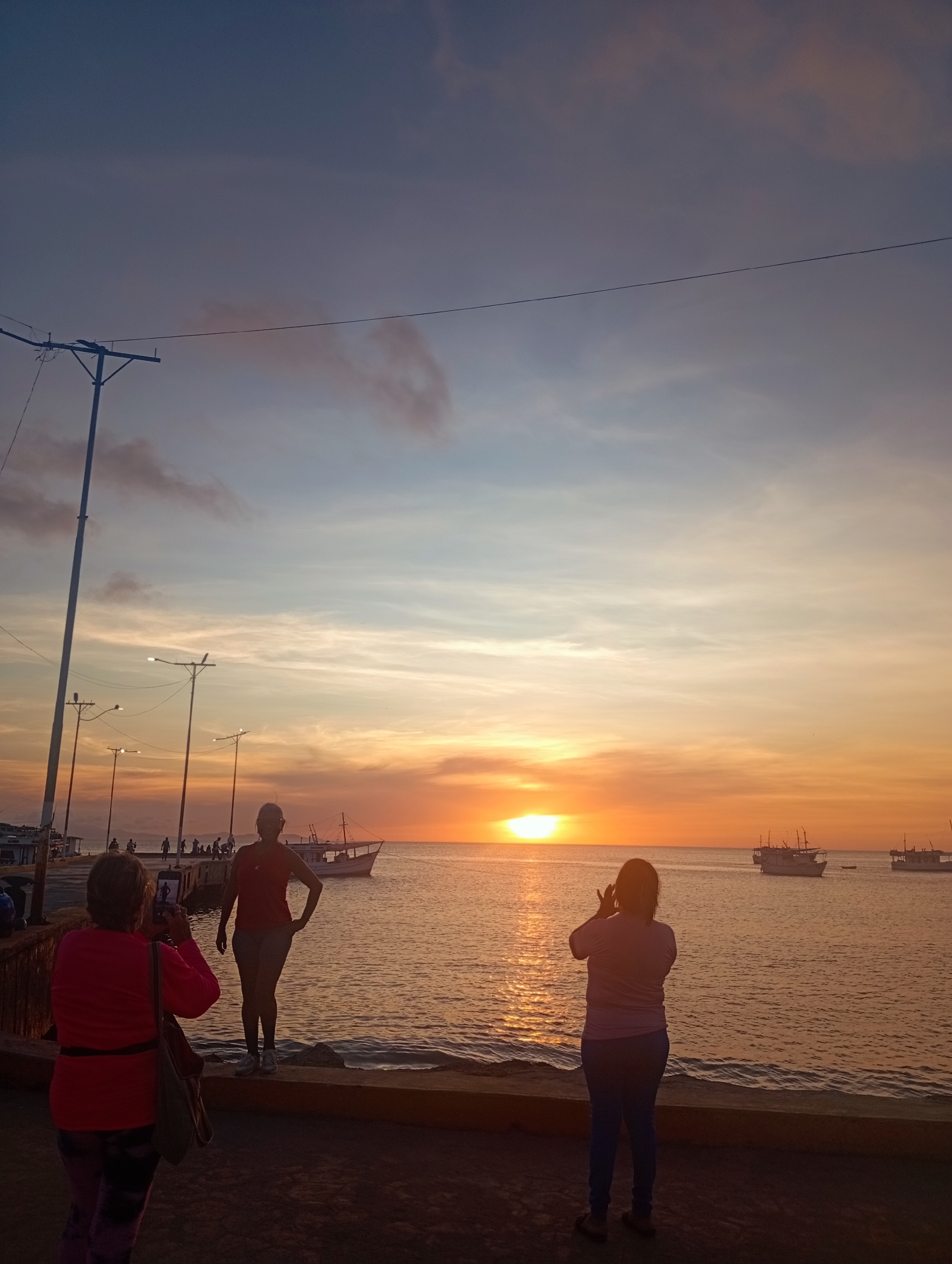 The image captures a sunset scene in Juan Griego, Venezuela. The sun is setting over the ocean, casting a warm, orange glow across the water and sky. In the foreground, three figures are visible. On the left, an individual in a pink shirt is holding a phone, seemingly taking a picture. In the center, a person in a red top and shorts stands on the edge of the concrete barrier, posing. On the right, another figure in a light-colored shirt and blue jeans also holds a phone, possibly taking a photo. Boats are anchored in the distance. Utility poles and streetlights line the walkway along the water's edge.