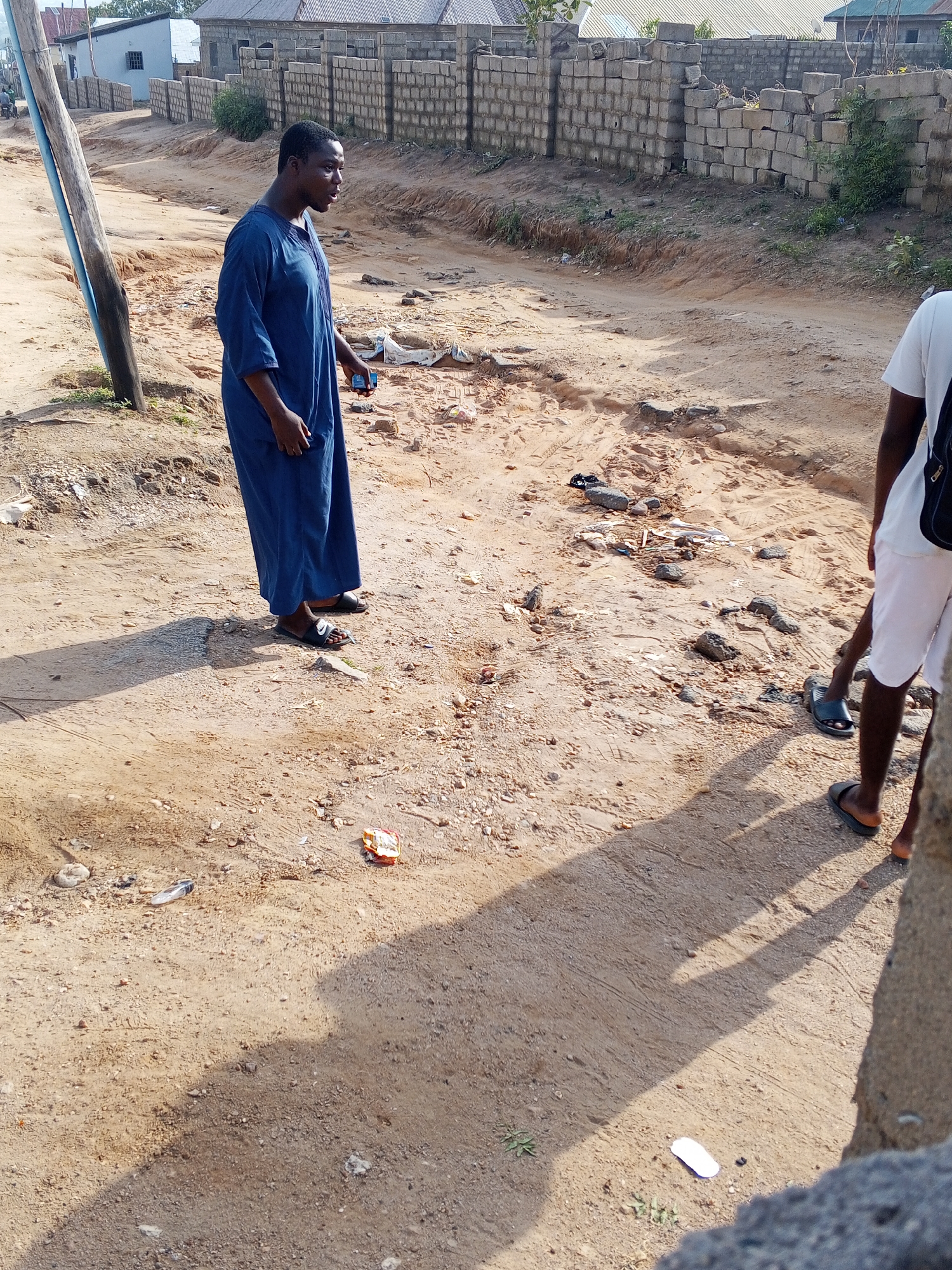 The image shows a dirt road in Adonkolo, Nigeria. A man in a blue robe stands on the left side of the road, holding a phone or small object. Another person, partially visible, wearing a white shirt and shorts, stands on the right. The road surface is uneven and contains debris. A concrete block wall lines the side of the road. A utility pole is on the left. Several buildings are visible in the background.