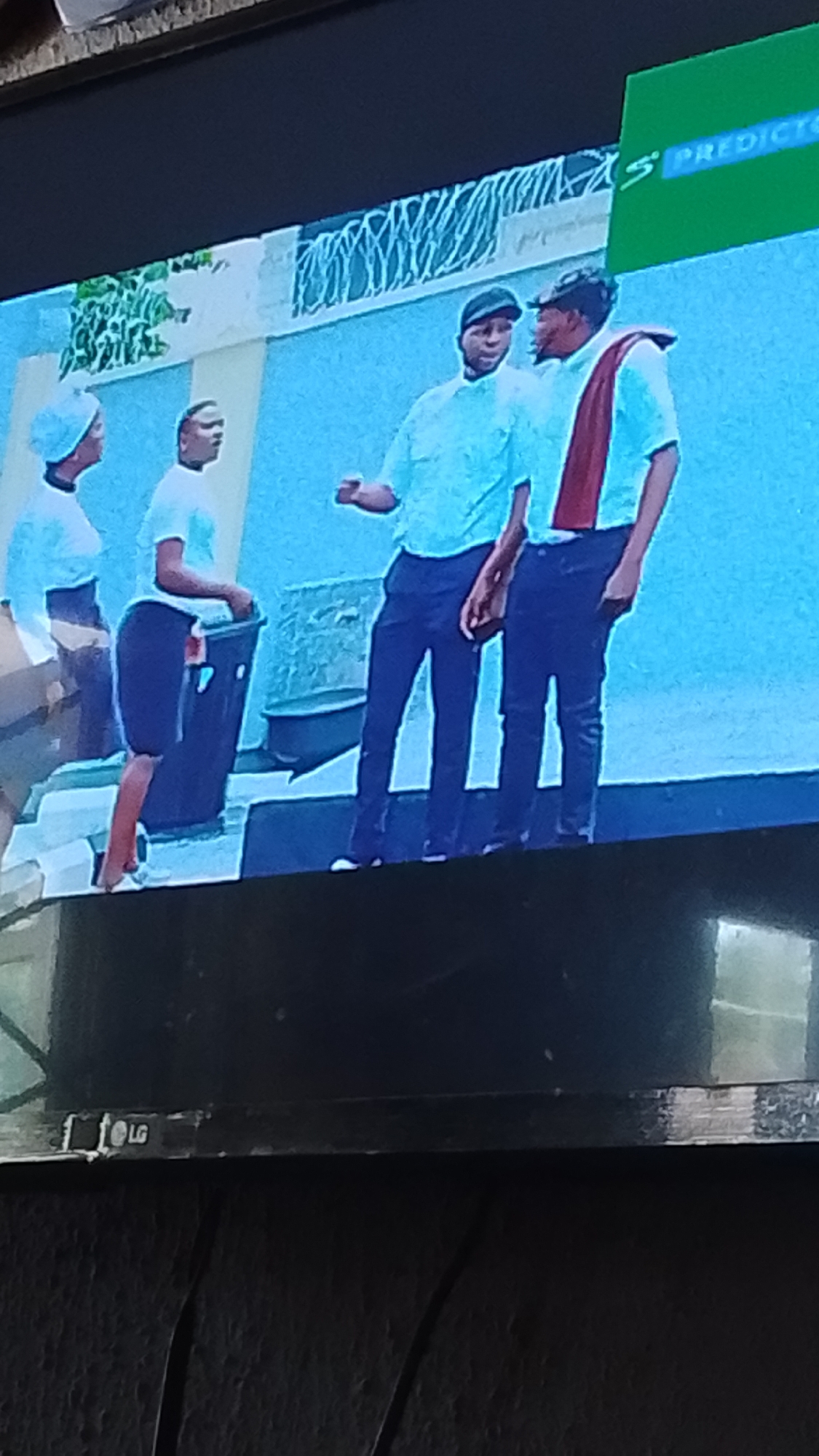 In an urban scene in Lokoja, Nigeria, three individuals are captured on a television screen. On the left, a woman wearing a white head covering and a white top stands, facing right. Next to her, another woman with a white top is seen from the side, bending slightly to interact with a dark waste bin. In the center and right of the frame, two men, both wearing light-colored shirts and dark trousers, are engaged in conversation. One man, on the left, wears a dark cap and gestures with his right hand. The other man, on the right, has a dark cap and a maroon sash draped over his left shoulder and across his chest. The background features a light-colored wall with a decorative element resembling a fence, and a vibrant green banner with white text, partially visible at the top right of the screen, displaying "PREDICTO". Below the television screen, the "LG" logo is visible on the bezel.