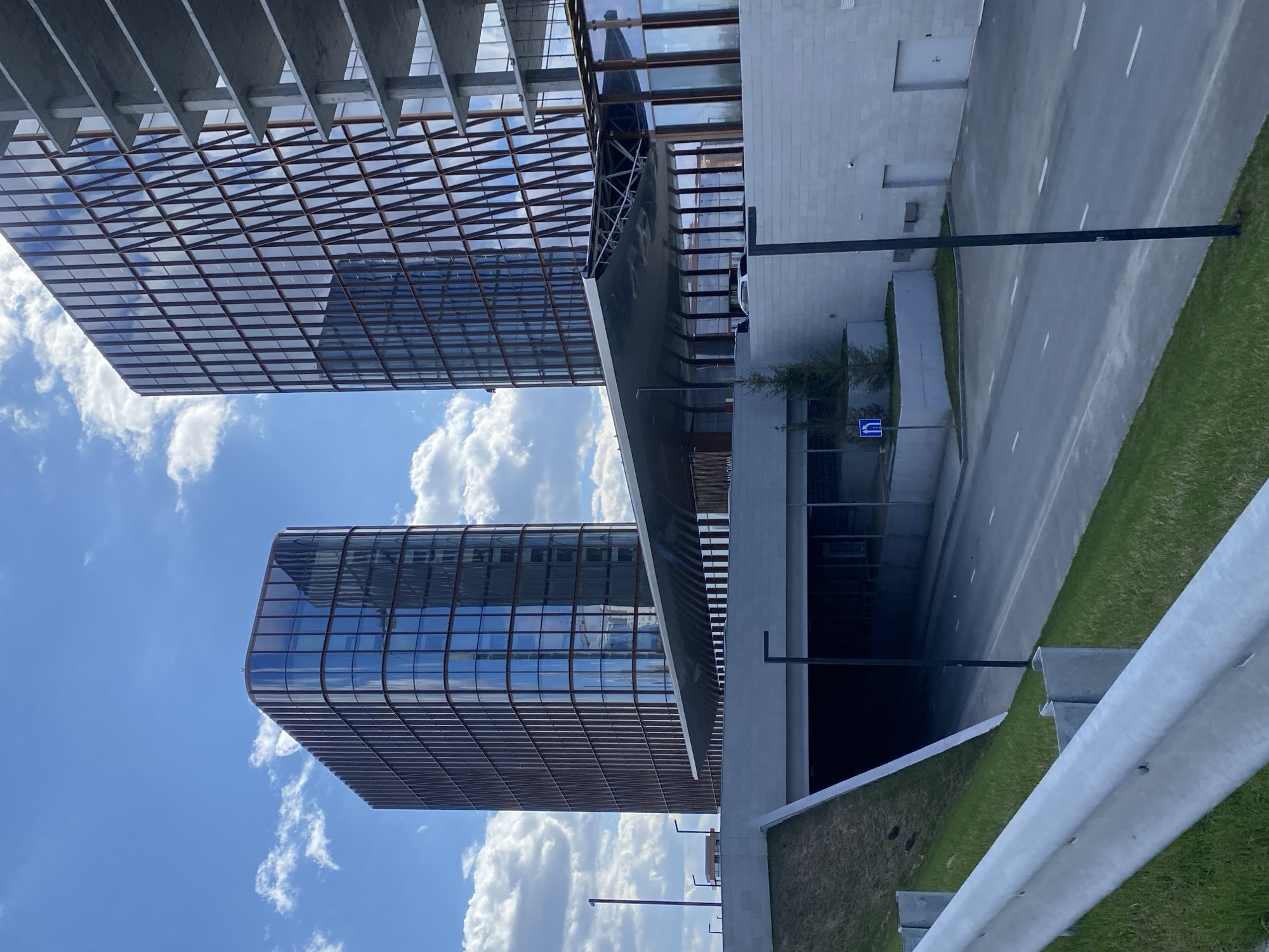 The image shows a view in Kazan, Russia, focusing on modern architecture and infrastructure. Two buildings with curved facades and glass windows are visible against a blue sky with scattered clouds. An overhead structure with a grid-like design partially obscures the top building. A roadway with white lane markings runs alongside one of the buildings, bordered by grass and a safety barrier. A pedestrian crossing sign is visible near the road.