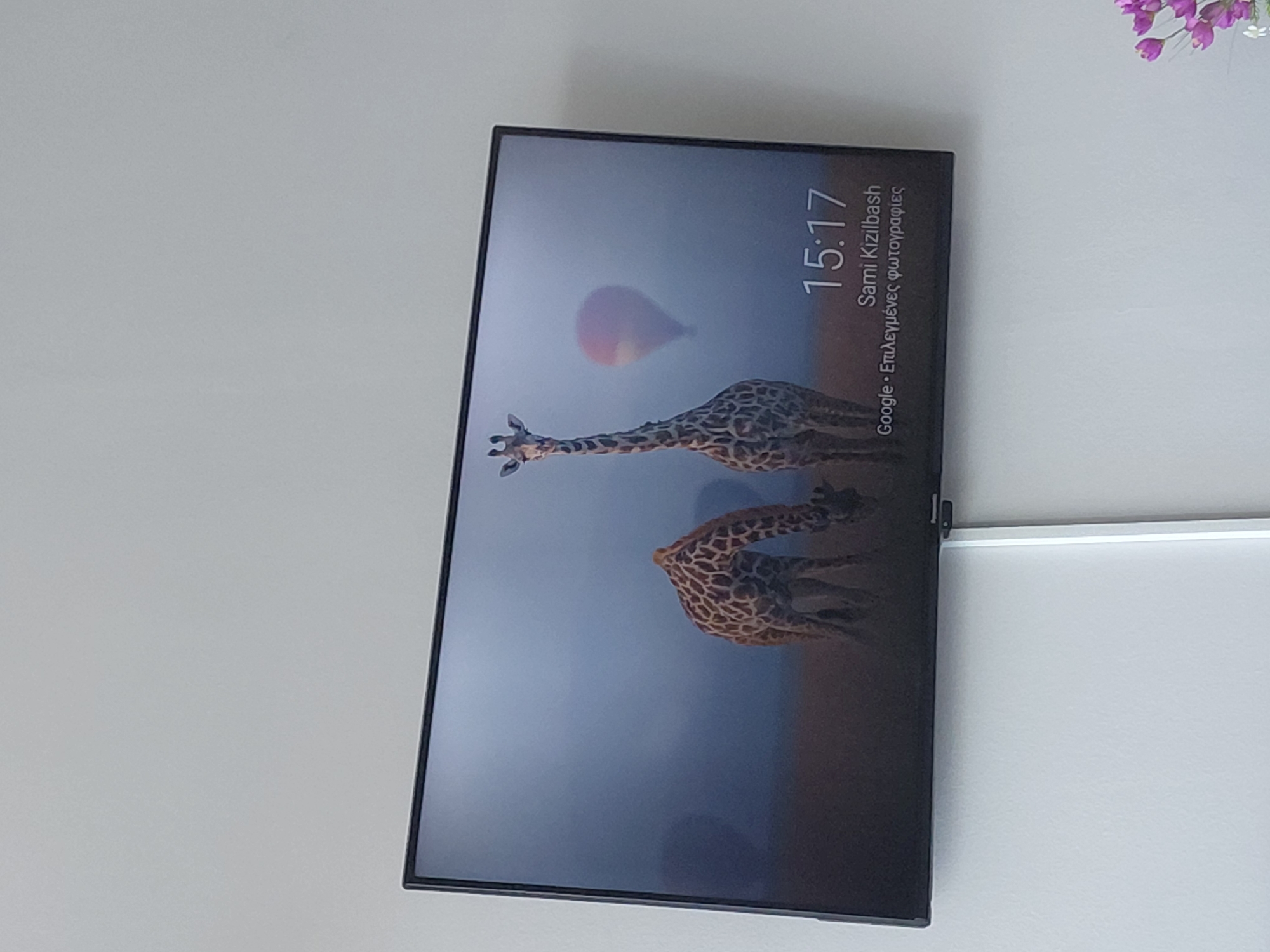 The image shows a television screen displaying a scenic image of two giraffes in a misty landscape, with a hot air balloon in the background. The time on the screen reads 15:17, and the text below indicates "Sami Kizilbash" and "Google · Επιλεγμένες φωτογραφίες" which translates to "Google · Selected photos" in Greek. The television is mounted on a light-colored wall, likely in a domestic setting. The lower right corner of the image shows a hint of purple flowers. There is no specific information in the image to indicate a location within the Municipal Unit of Aegio, Greece.