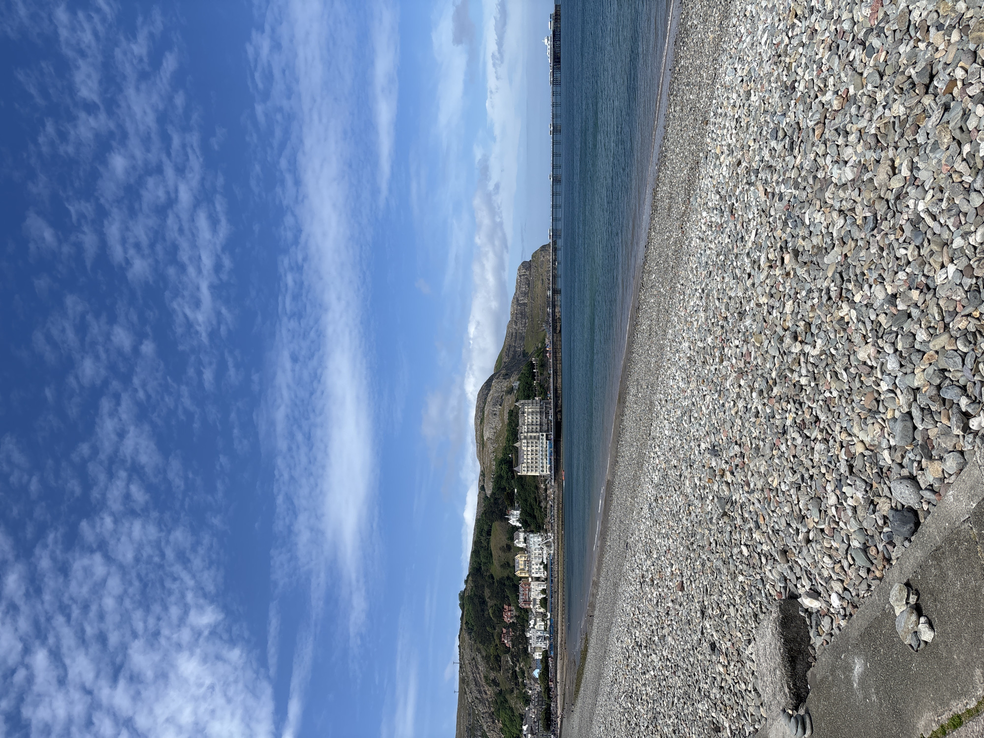 The image shows a pebble beach in Llandudno, United Kingdom, under a blue sky with scattered clouds. The beach is composed of small, gray pebbles. In the distance, a mountain slopes down towards the coastline, with buildings situated along its base and on the lower slopes. A pier extends into the sea. The water appears calm. A concrete barrier runs along the edge of the beach in the foreground.
