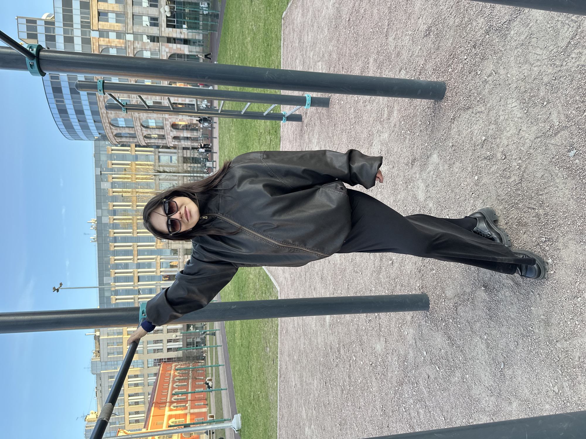 An outdoor, vertically oriented (when corrected) full-shot photograph depicts a young woman posing next to calisthenics bars in an urban park or sports ground in Saint Petersburg, Russia.
The woman, with long dark hair, is positioned with her body angled towards the camera, leaning slightly to her left. She wears dark sunglasses, a black leather-look bomber jacket, black wide-leg trousers, and chunky black platform shoes. Her right hand grasps a dark gray horizontal metal bar, while her left arm is slightly bent at her side. She looks directly at the camera with a neutral expression, possibly a faint smile.
The ground around her is covered in light-colored gravel or small pebbles. Several dark gray horizontal metal bars, connected by light blue/green plastic clamps, form what appears to be outdoor exercise equipment. Beyond the gravel, a strip of well-maintained green grass is visible. The background features a clear blue sky and a mix of urban architecture: modern multi-story buildings with reflective glass facades stand alongside older, more traditional buildings constructed from brick or stone, characterized by arched windows and ornate details. The bright, clear daylight suggests the photo was taken during late morning or afternoon. No discernible text is visible in the image.