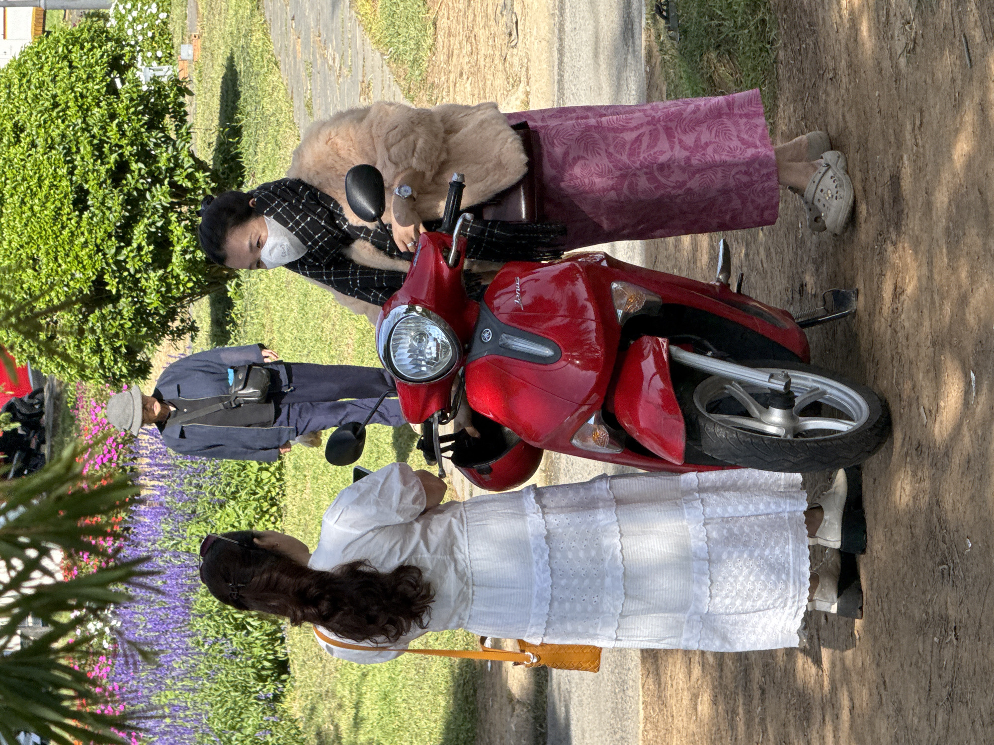 In Da Lat, Vietnam, two women stand near a red scooter. The woman on the left wears a white dress and carries an orange bag. The woman on the right wears a patterned pink skirt and a light brown fur jacket, with a black and white scarf and a face mask. She is wearing clogs. In the background, a person in a hat and blue jacket walks past a vibrant display of purple and pink flowers, with a lush green bush behind them. The ground is a mix of dirt and paved pathways.