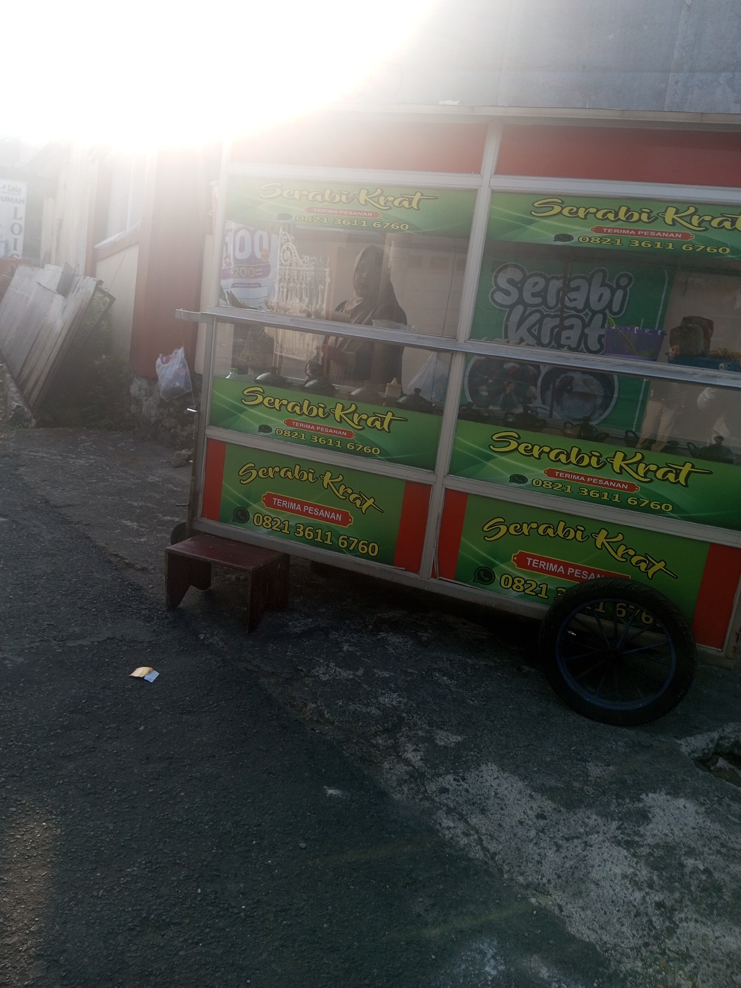 A mobile food cart, identified by the signage "Serabi Krat" and the phone number "0821 3611 6760", is parked on an asphalt surface. The cart features a glass display case and is decorated with green panels bearing the business name and a phone number. A small, dark-colored stool is placed in front of the cart. The background shows a building with sunlight casting a bright glare, partially obscuring details of the environment, which appears to be an outdoor urban setting in Krasak, Indonesia. There are visible people inside the cart's display area, and a wheel is attached to the cart. Debris is scattered on the ground in front of the cart.