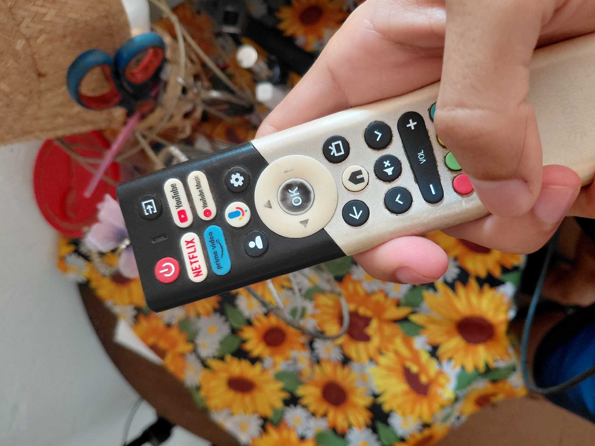 A right hand, belonging to an individual with light skin, holds a two-toned smart television remote control in a domestic indoor setting. The remote has a black upper panel and a beige lower body, showing signs of use and grime, particularly around the central "OK" button. The thumb is positioned over the "VOL -" section of the volume control bar.
The remote features a red power button, dedicated quick-access buttons for NETFLIX (red), prime video (blue), YouTube (red with play icon), and YouTube Music (red with play icon). Other visible buttons include a microphone icon (likely for voice control), a gear icon (settings), a person icon (profile/account), a central circular navigation pad with an "OK" button, directional arrows, and various function buttons for back, home, input, mute, and confirmation (checkmark). Four small, colored (red, green, blue, yellow) function buttons are partially visible at the bottom.
The background is predominantly covered by a fabric displaying a vibrant pattern of yellow and orange sunflowers with dark brown centers, interspersed with white daisy-like flowers and green leaves on a dark (likely black) base. In the upper right background, a blurred woven basket or container is visible, holding a pair of blue-handled scissors with orange blades, a pink drinking straw, and a red plastic cup or container. The upper left background shows a blurred, light-colored surface. A dark cable runs diagonally across the mid-left background. The bottom left shows a glimpse of a blue object. The exact location in the city of Unknown, Unknown is not discernible from the image.