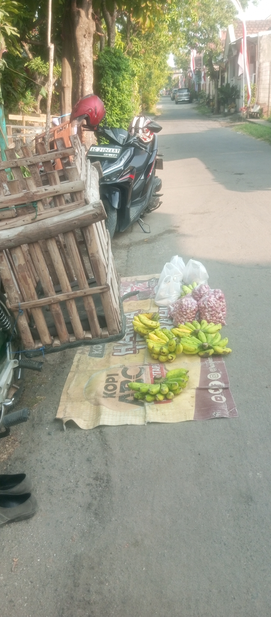 A street scene in Kediri City, Indonesia, shows a red helmet resting on a motorcycle. To the left of the motorcycle is a wooden cage, possibly for livestock. In front of the cage and motorcycle, on a patterned mat on the asphalt road, are several bunches of bananas and bags of what appear to be onions. The street is lined with houses and trees. In the background, more houses and a few parked cars are visible. The image is taken from a slightly elevated angle, looking down the street.