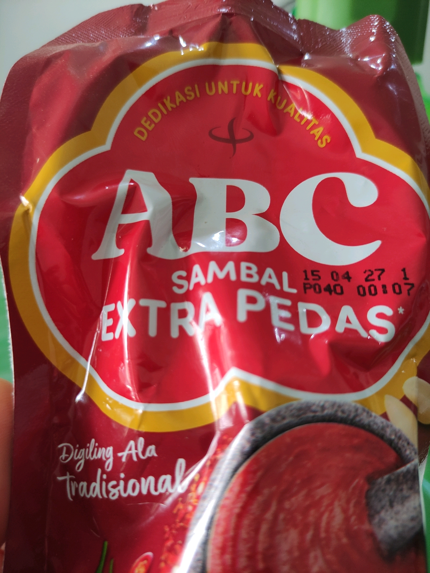 The image shows a close-up of a red pouch of ABC brand chili sauce. The pouch features a large white "ABC" logo with a yellow and white border. Text on the pouch reads "DEDIKASI UNTUK KUALITAS" and "SAMBAL EXTRA PEDAS". There are also date markings visible: "15 04 27 1" and "P040 00:07". Below the main logo, in smaller white cursive text, it says "Digiling Ala Tradisional", which translates to "Ground in a Traditional Way". The bottom of the pouch shows a graphic of chili peppers and what appears to be a mortar and pestle, with the sauce inside. The background is out of focus and appears to be a light-colored surface, possibly a table, with some green elements also out of focus. The image is taken from a slightly downward angle. No specific location in Bulukumba, Indonesia can be determined from the image.