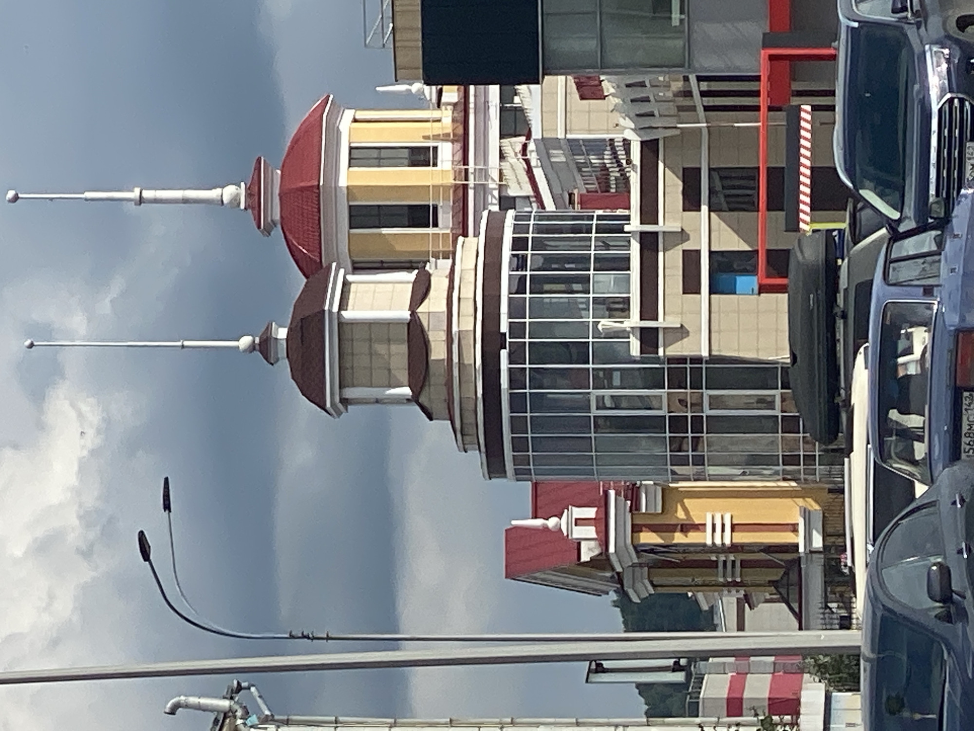 The image shows a contemporary architectural scene in Novokuznetsk, Russia, featuring a cluster of modern buildings with distinctive red roofs and ornate spires. A tall street lamp with two lights stands to the left, silhouetted against a cloudy, grey sky. In the foreground, several cars are parked, including a dark blue sedan and a dark grey SUV, partially obscuring the base of the buildings. The buildings themselves exhibit a mix of architectural styles, with prominent glass facades on a cylindrical structure and decorative elements on other sections, such as yellow accents and white trim. The overall impression is one of urban development with a touch of decorative flair.