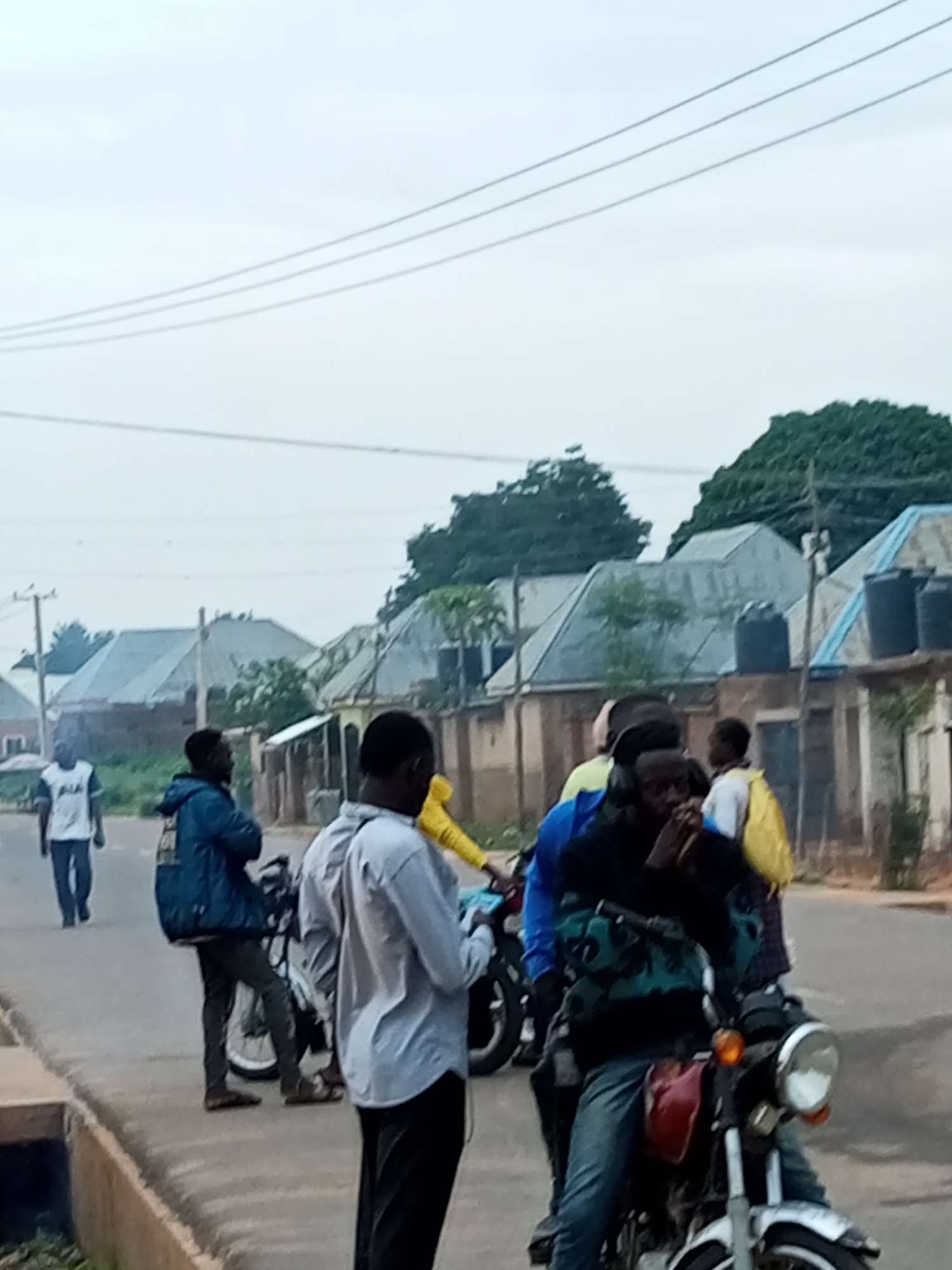 A group of people are gathered on a street in Tempure, Nigeria, under an overcast sky. In the foreground, a man wearing a dark jacket over a patterned sweater and jeans is seated on a red motorcycle. To his right, another person in a light grey shirt and dark pants stands, looking towards the motorcycle. Behind the motorcycle rider, another individual in a bright yellow jacket is partially visible. To the left of the main motorcycle, a person wearing a blue jacket and dark pants stands next to a motorcycle. Further down the street and to the left, a lone figure in a white t-shirt and dark pants walks away from the camera. In the background, several buildings with corrugated metal roofs and blue water tanks are visible, with trees and utility poles interspersed. The scene appears to be a residential or mixed-use area.