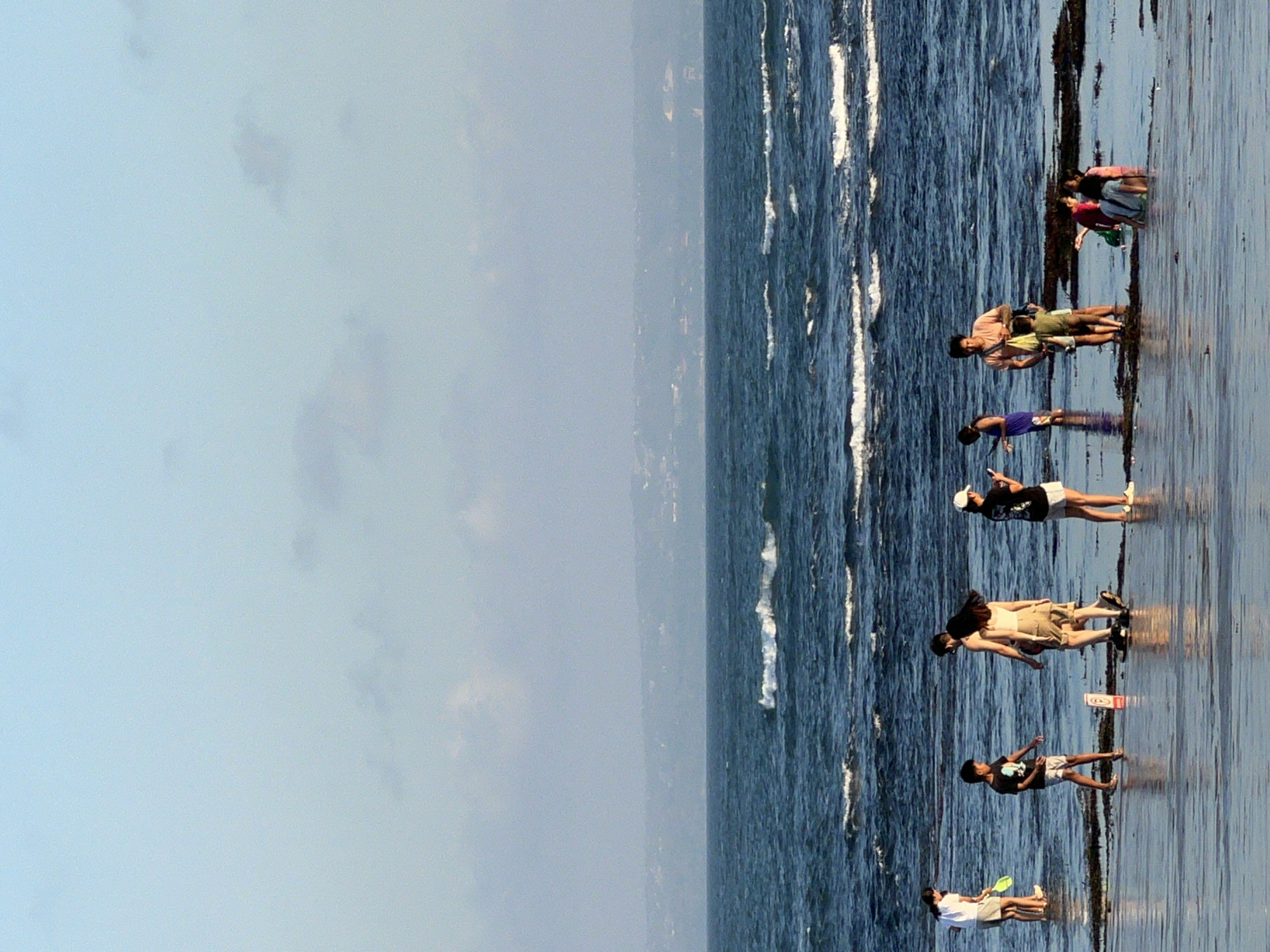 A vertical, eye-level medium shot captures a group of people on a shallow, wet beach with calm water and distant waves under a bright, partly cloudy sky. The sand is dark and wet, with patches of seaweed. In the mid-ground, a line of people strolls near the water's edge. On the left, a woman in a white t-shirt and shorts holds a green paddle. Next to her, a boy in a black t-shirt and shorts stands and gestures. Behind them, a couple walks, the man in a light pink t-shirt and khaki shorts, and the woman in a white tank top and khaki shorts. To their right, a woman in a black t-shirt and shorts holds a phone. Further right, a man in a light pink t-shirt and shorts stands with a yellow bag over his shoulder, facing away from the camera, while two young children in green and red shorts stand in front of him. In the shallow water to the far right, a group of children, one in a pink swimsuit, are visible. The background features a wide expanse of dark blue ocean with small waves breaking. In the far distance, a hazy coastline with low hills and some buildings is visible. The scene is likely in North Kuta, Indonesia, given the beach setting and distant landscape.