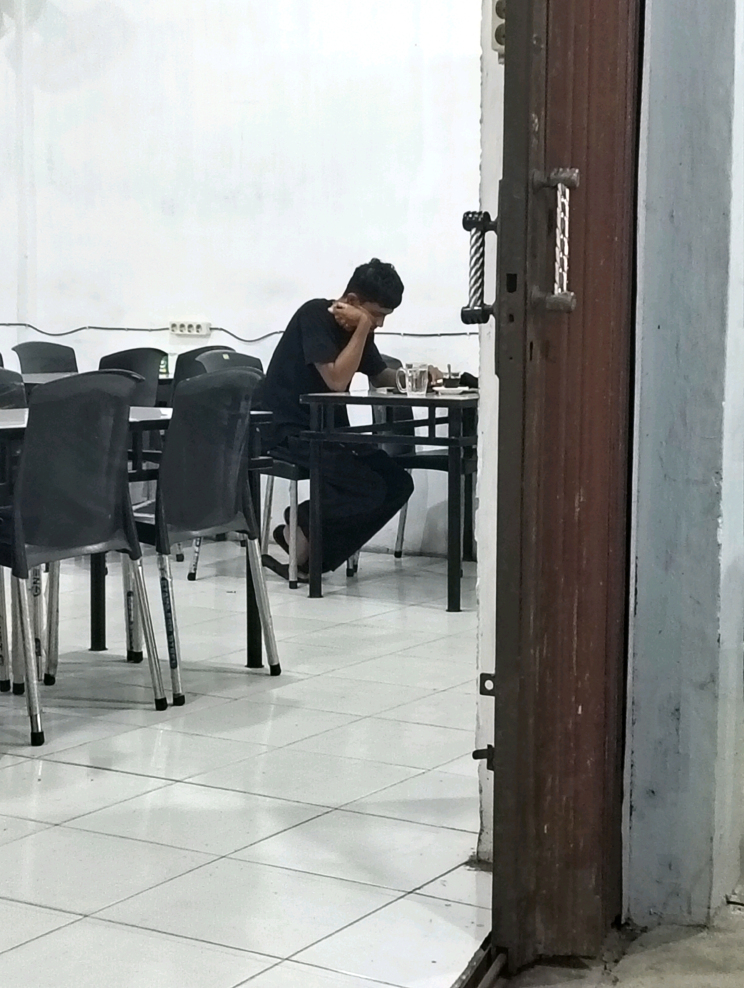 A young person, appearing to be male, is seated at a table in what appears to be a dining establishment. The individual is wearing a black t-shirt and dark pants. Their head is resting on their hand, and they are looking down at the table, suggesting a posture of thought or weariness. On the table in front of them are a glass of water and what looks like food on a plate. The environment is a room with white walls and a tiled floor. Several empty black chairs are visible, arranged around tables. A prominent wooden door with metal handles is situated on the right side of the frame, partially obscuring the view into the room. The overall impression is of a quiet moment, possibly during off-peak hours, in a humble eatery in Grong-Grong, Indonesia.