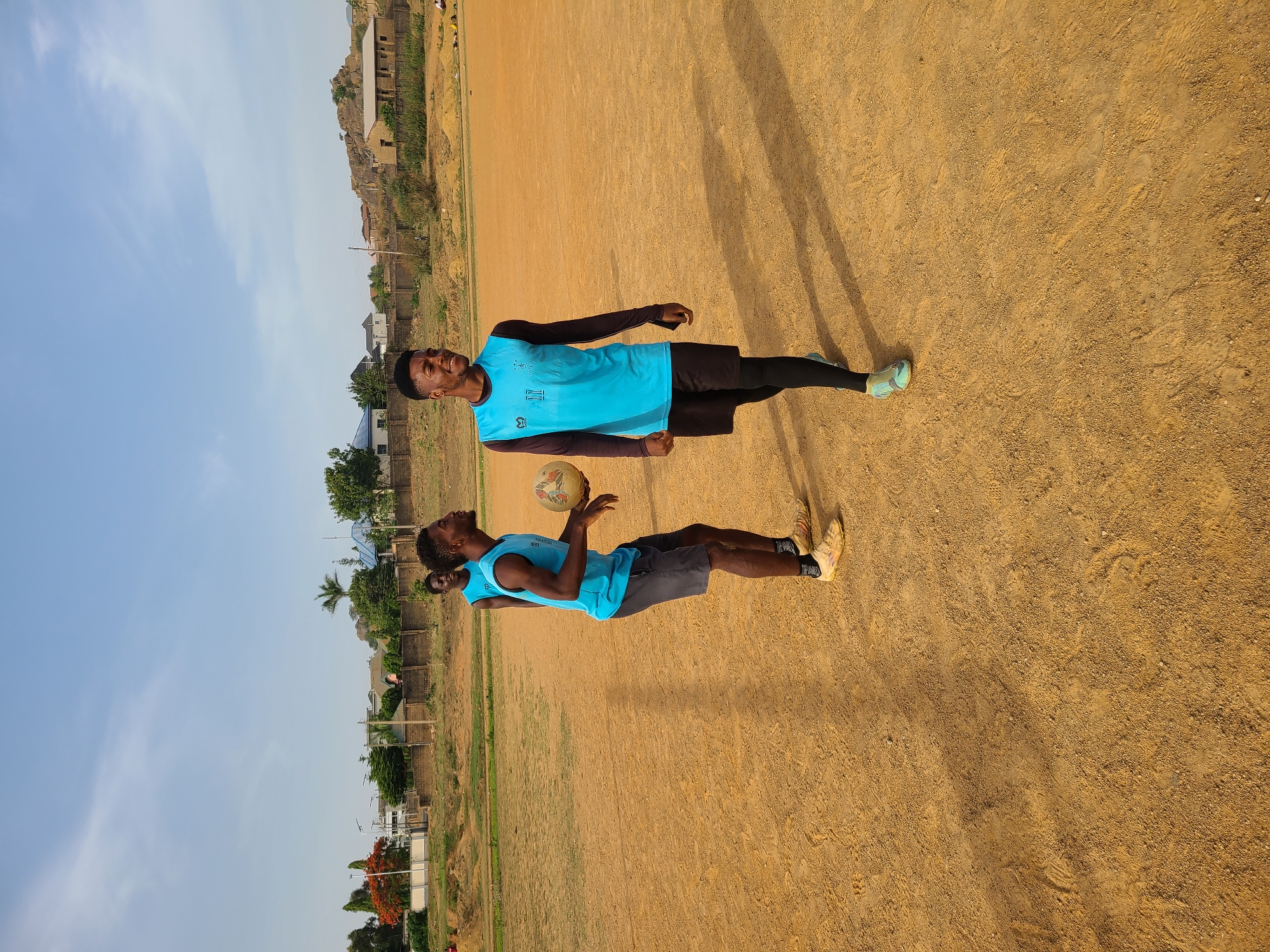 Two men are standing on a dirt field in Barikin Jirga Summa, Nigeria. One man is holding a soccer ball and wearing a light blue jersey and grey shorts with tan sneakers. The other man is wearing a light blue jersey with black leggings and black shorts, and mint green sneakers. The background includes grass and a building on a hill in the distance under a clear blue sky.