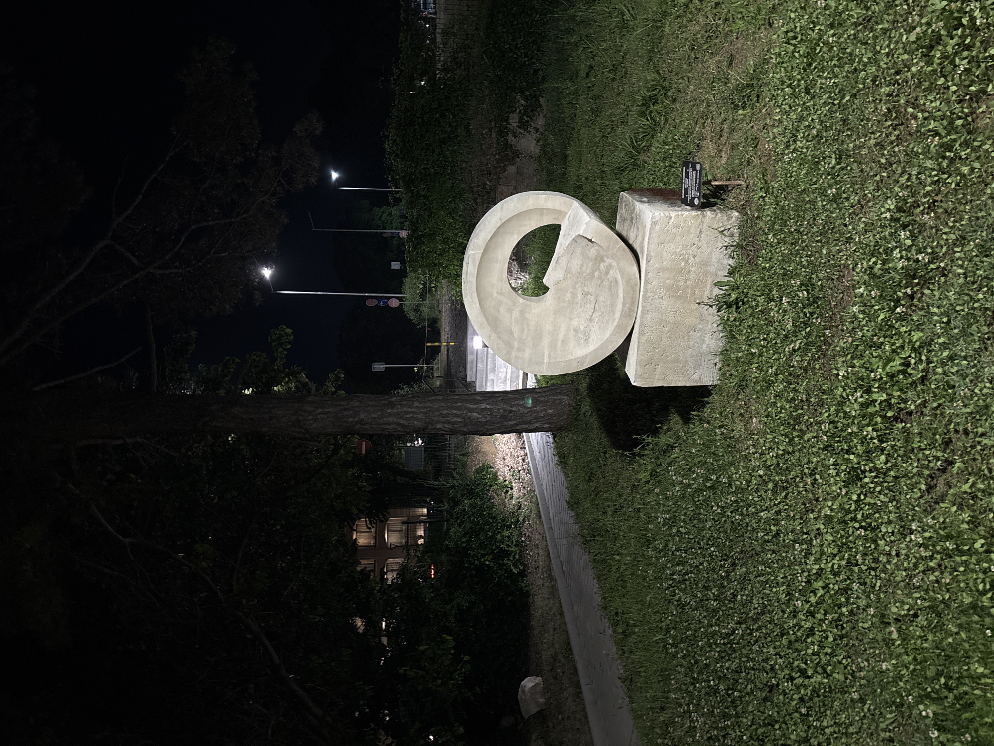 The image depicts an outdoor scene at night, illuminated by artificial lighting from street lamps and architectural lights. In the foreground, a circular stone sculpture is prominently displayed on a concrete base, situated on a grassy area with scattered clover. To the left, a paved walkway leads towards a set of stairs. Further back, trees and dense vegetation form a backdrop, with a building visible in the distance. Street lights are positioned along what appears to be a road. The location is identified as the Sea Garden in Varna, Bulgaria. There are no people or animals visible in the frame.