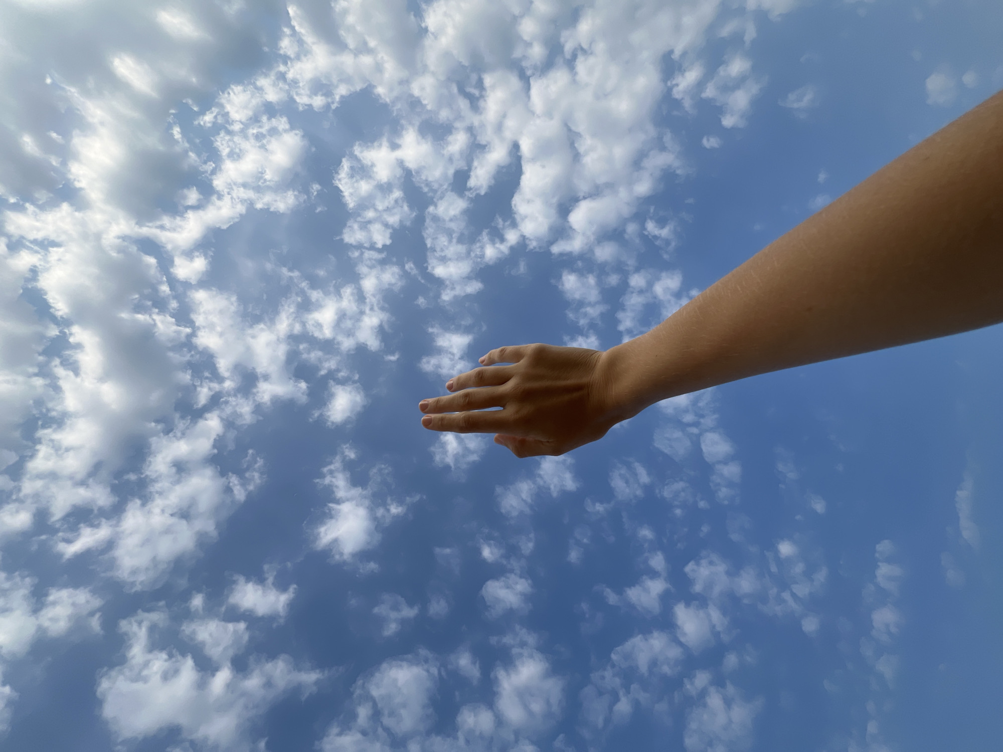 A low-angle shot captures a person's bare, light-skinned arm and hand extending diagonally from the upper right corner towards the center-left of the frame. The arm appears outstretched or reaching upward, with the hand slightly open and fingers extended. The thumb and index finger are visible, pointing generally forward.
The background is a vast, clear blue sky, predominantly bright. It is filled with numerous scattered white and light gray clouds, ranging from fluffy cumulus formations to more wispy structures. The bright illumination on both the clouds and the arm suggests it is daytime, likely a sunny day with some cloud cover. The overall scene creates a sense of openness and expanse, with the human element interacting with the natural environment.
No specific activity beyond the gesture of the hand is discernible. There are no other visible people, objects, or text within the image. While the image itself lacks direct visual cues for a location, the provided context indicates that the media file is from Nam Nha Trang, Vietnam. The focus of the image is the interplay between the human form and the sky, emphasizing a connection to the vast outdoor environment.