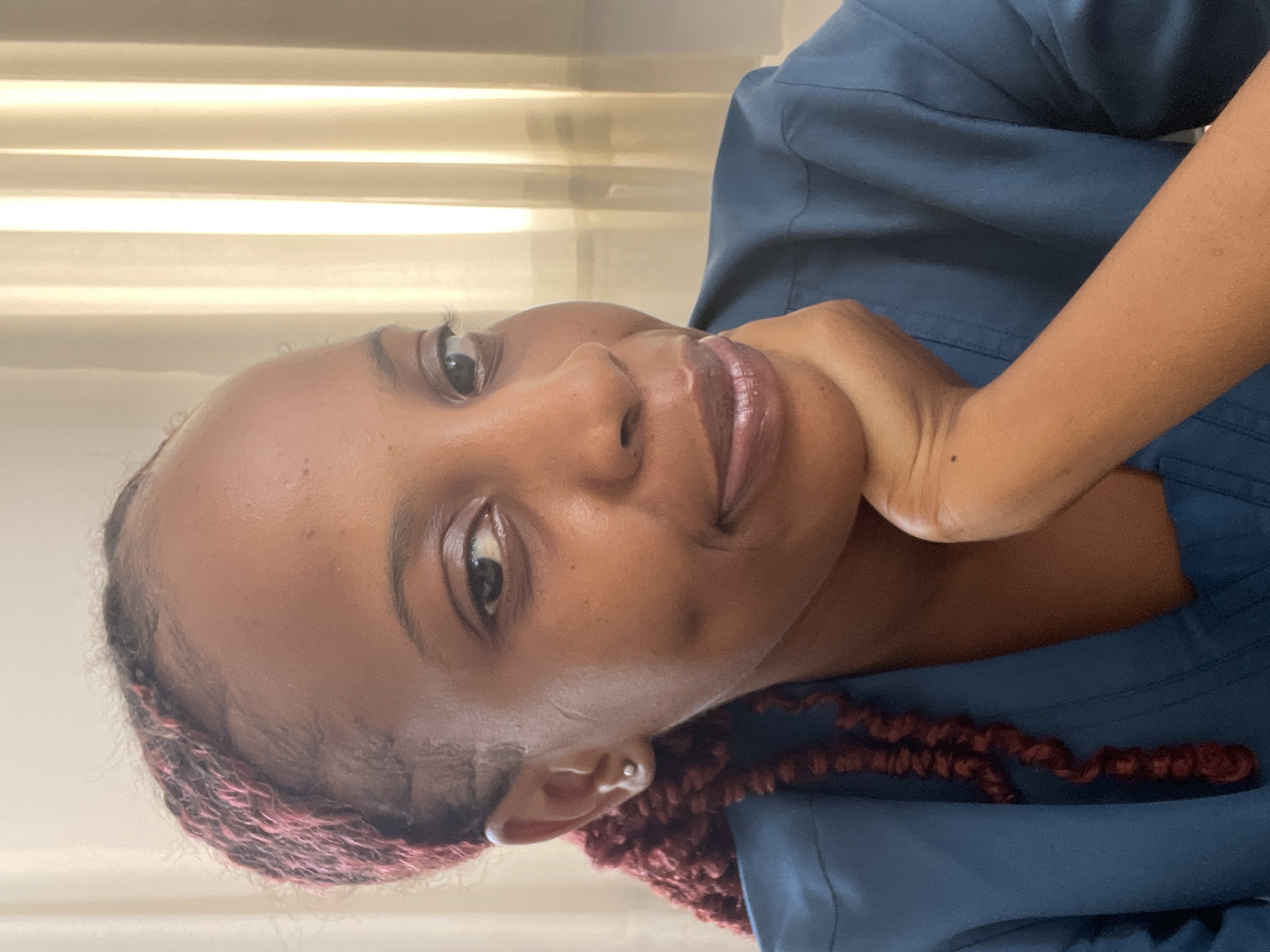A medium shot shows a Black woman with braided dark hair with red streaks, wearing a blue top. She has dimples and is resting her chin on her hand, looking slightly to the right. The background is a blurred curtain with vertical folds. The image appears to have been taken indoors.