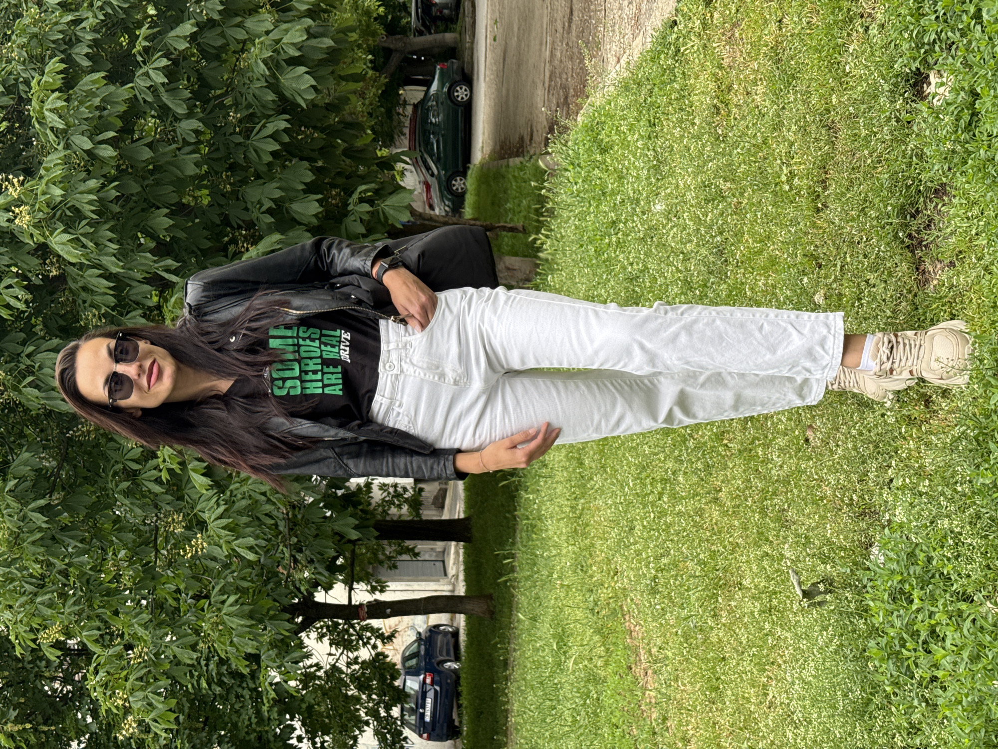 A young woman stands on a grassy lawn near trees. She wears sunglasses, a black t-shirt with green text, a black leather jacket, white jeans, and tan sneakers with white socks. She faces the camera with her left hand in her pocket and her right hand near her hip. Behind her are trees and a glimpse of cars parked on a street, suggesting a park-like setting in Pleven, Bulgaria.