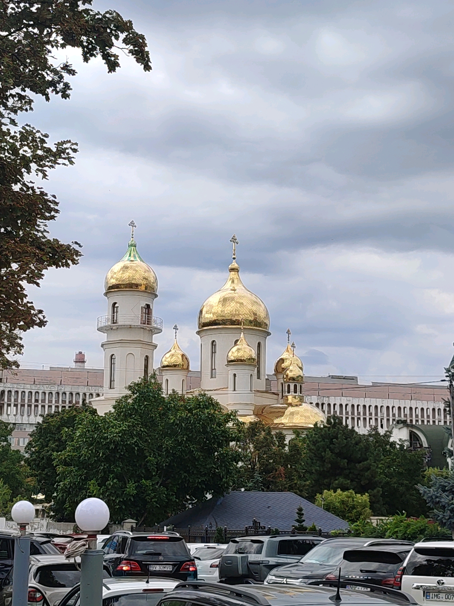 The image depicts the Nativity Cathedral in Chișinău, Moldova, a prominent landmark with its striking golden domes. The cathedral is partially obscured by lush green trees in the foreground, with a dense cluster of cars parked in front, suggesting a public space or thoroughfare. The sky is overcast with heavy, grey clouds, hinting at possibly inclement weather or a late afternoon scene.
Behind the cathedral, a large, multi-story building with arched windows stretches across the frame, possibly an administrative or residential structure. The overall impression is one of an urban setting with a mix of religious and secular architecture, framed by natural elements. The presence of cars indicates that this is an accessible area, likely a street or parking lot in the vicinity of the cathedral. The lighting is diffused due to the cloudy sky, casting a soft, even light on the scene.