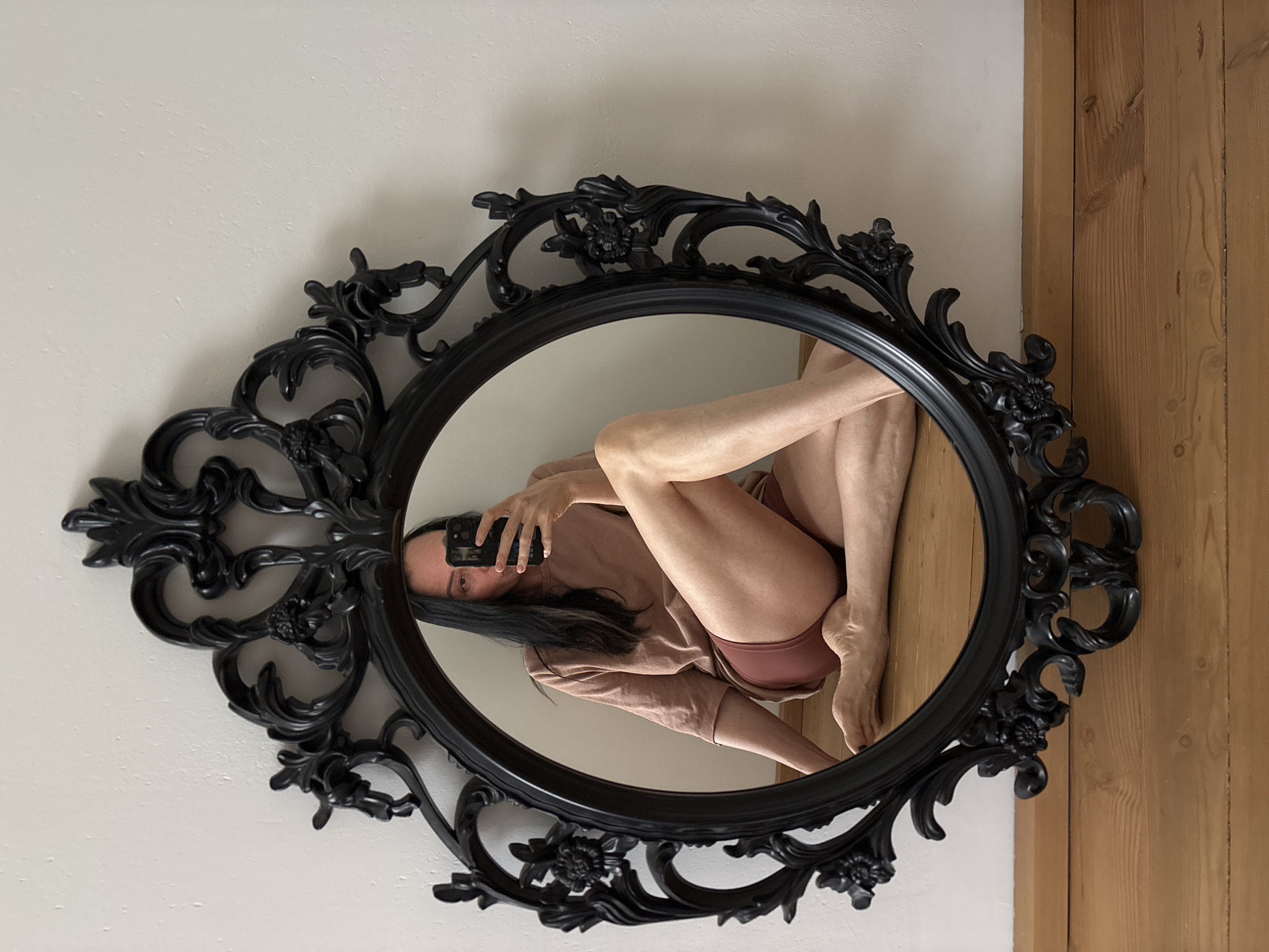 A framed oval mirror is mounted on a wall with white on the left and brown wooden planks on the right. A woman is reflected in the mirror, bent into an unusual position so her face, legs and midsection are all visible within the frame. She is holding a phone in her hand, presumably to take the picture. The environment suggests an interior setting, possibly a home. There is no contextual information to confirm the location in the city of Chaikovsky, Russia.
