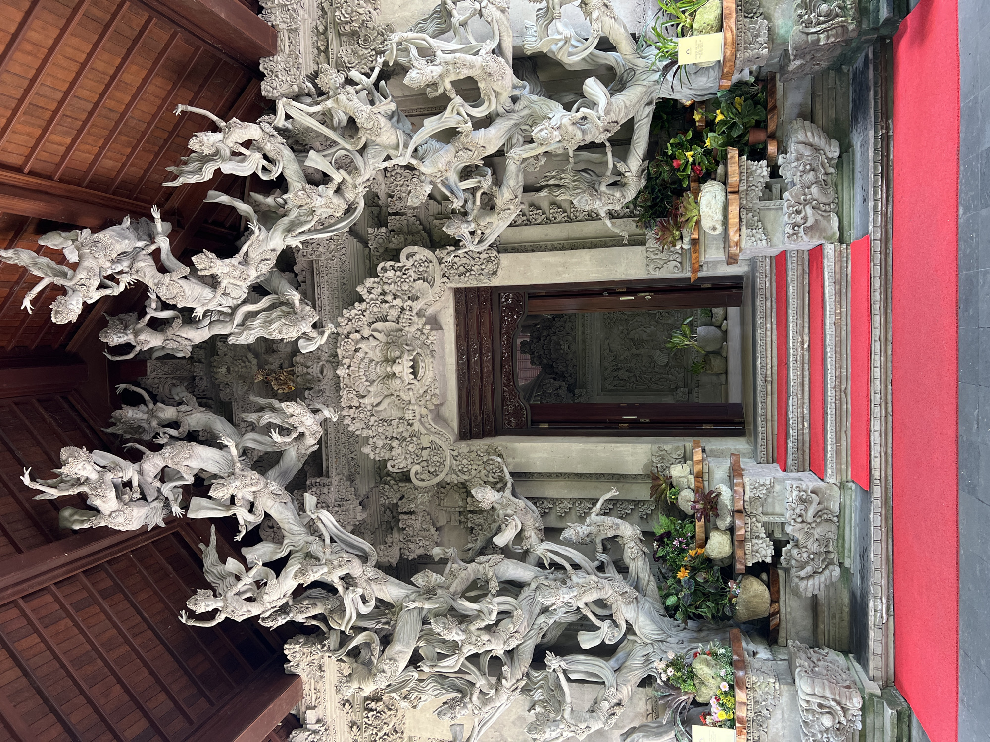 The photograph captures a detailed architectural scene in Kedewatan, Indonesia, displaying a building facade decorated with complex stone carvings. The carvings, in a light grey color, primarily depict human figures in graceful, flowing movements, possibly depicting dancers or mythological beings. These figures extend from the structure’s roof, constructed from dark brown wooden tiles.
Central to the composition is a doorway with a dark brown wooden frame. Flanking the doorway are stone structures, adorned with additional carvings and potted plants, which introduce color through green foliage and vibrant flowers. A red carpet covers a set of stairs, leading towards the door. The building’s walls are decorated with intricate carvings, adding depth and texture to the scene. The photograph is shot at a low angle.