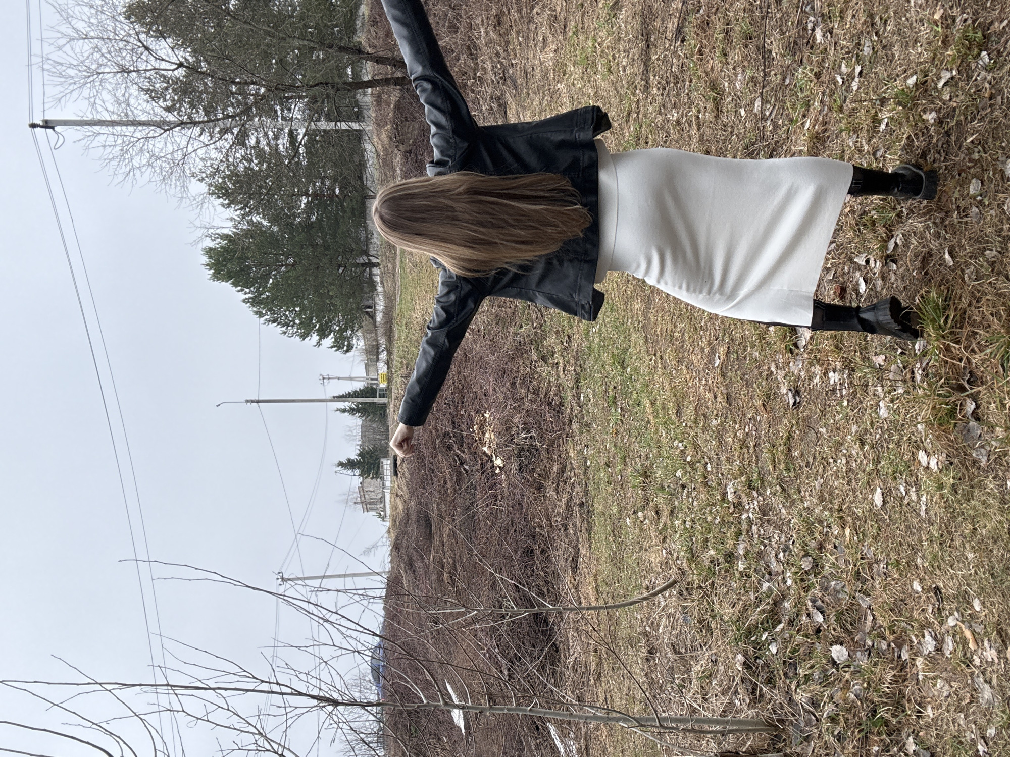 A woman stands in a field near Chaikovsky, Russia. She wears a black leather jacket, a white skirt, and black boots. Her arms are outstretched. The field is dry, with patches of green grass and fallen leaves. Trees and utility poles are visible in the background, under an overcast sky. Bare branches are prominent in the lower left corner.